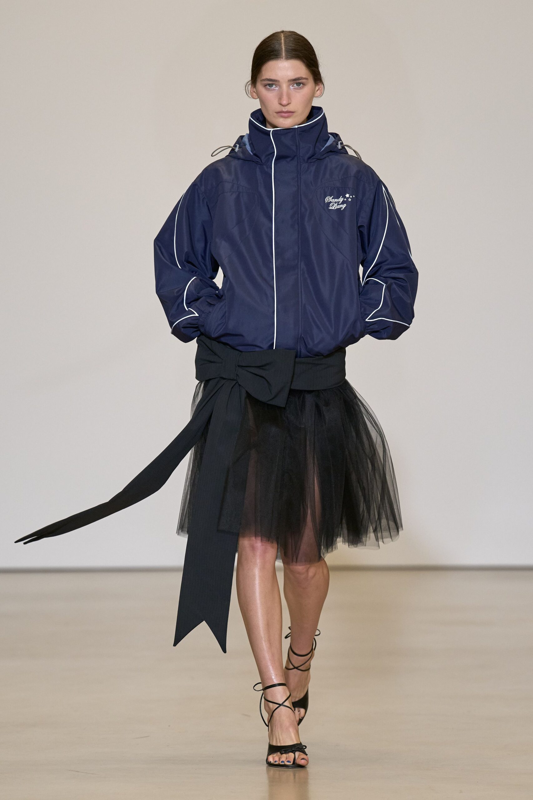 Navy nylon windbreaker with white piping and a high zipped collar. Wide black sash tied in an oversized bow, one tail dragging low. Black tulle skirt, sheer and softly belling from the hips. Black lace-up sandals, cords crossing the bare ankle. Walking out of a tennis club into dusk still wearing the recital skirt.