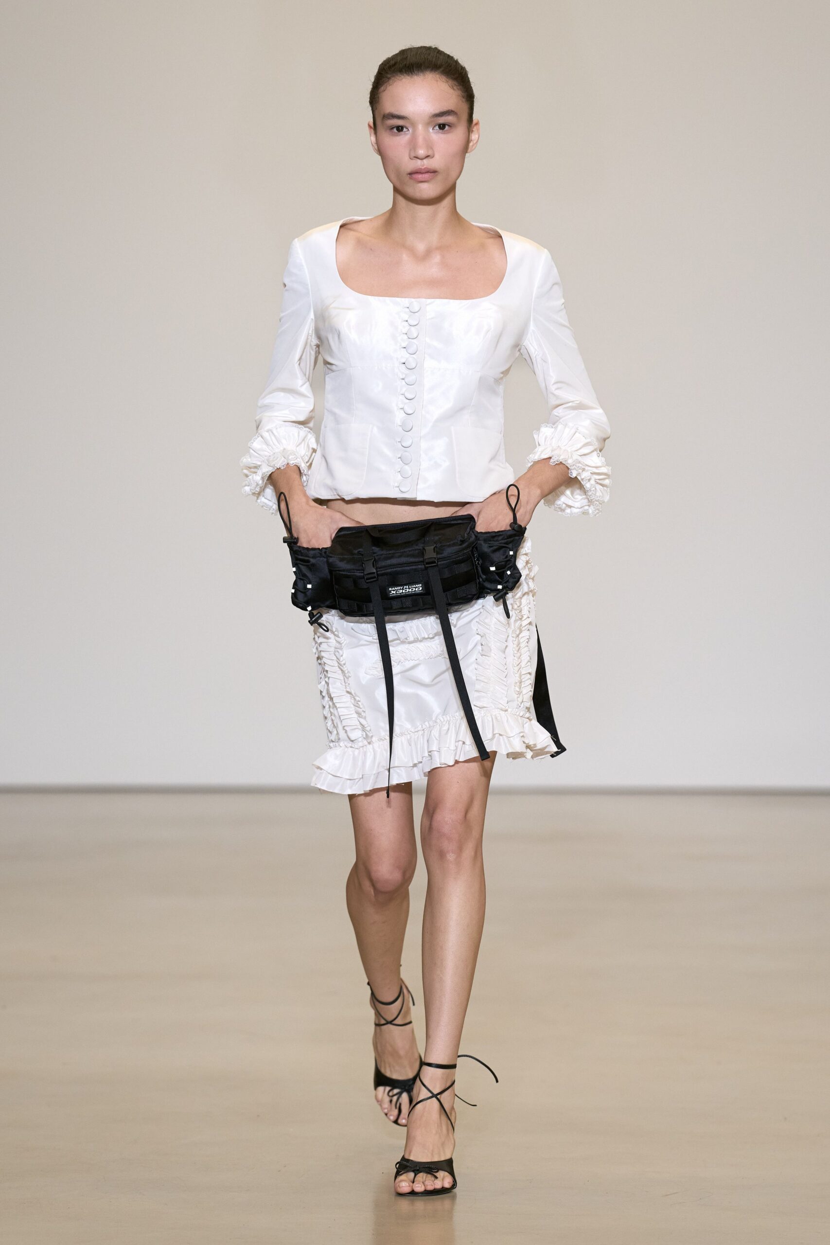 White satin blouse with a square neckline and self-covered buttons. Black nylon pouch belt, webbing straps hanging past the skirt. White satin mini skirt ruched into vertical ruffle channels. Black lace-up heeled sandals. For crossing a hotel lobby with room service menus still under your arm.