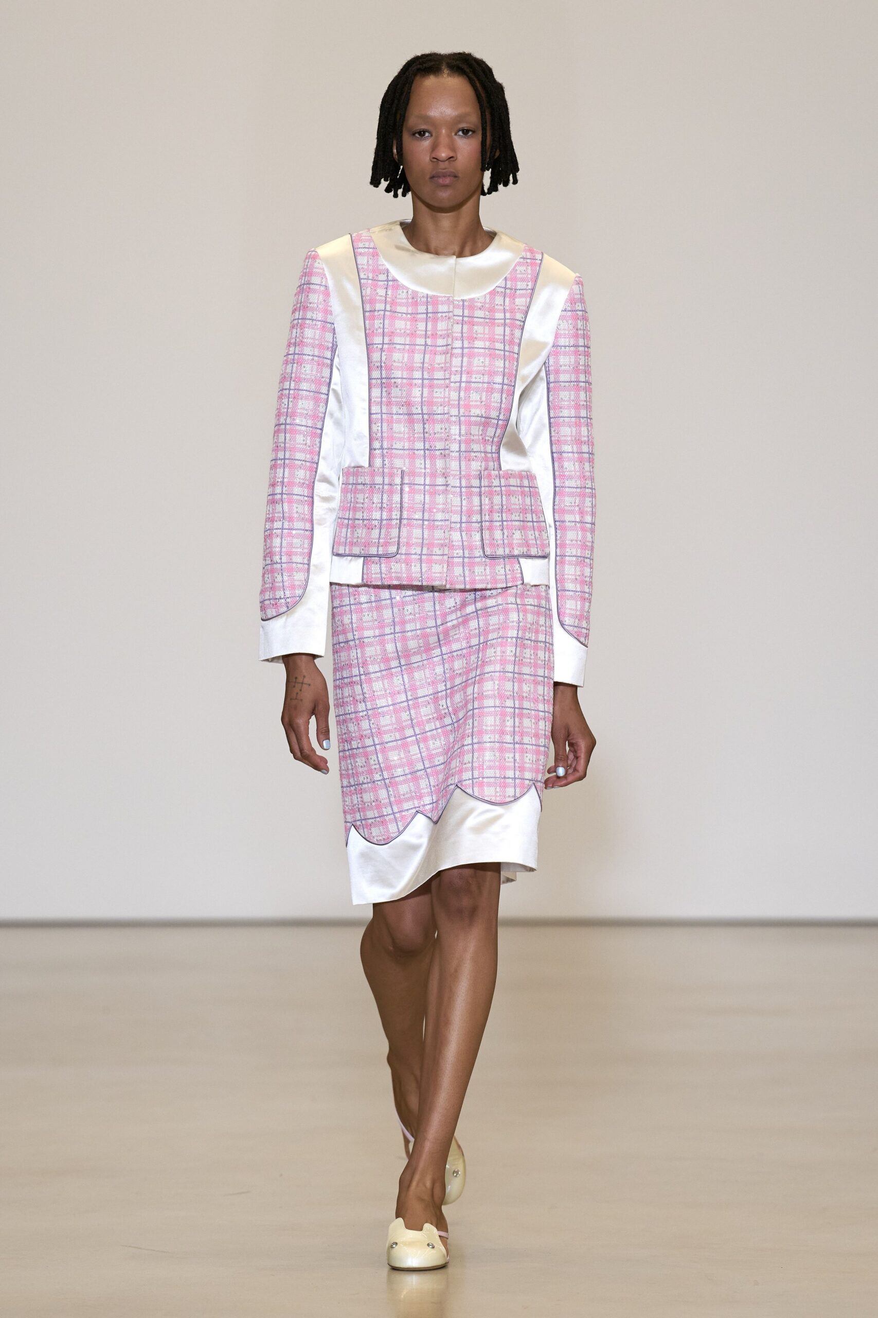 Pink windowpane tweed jacket with satin bib panels and rounded collar. Matching tweed skirt, scalloped hem lifting over a pearl-sheen underlayer. Cream satin mules with two tiny studs at the toe. Church-basement cake raffle, folding tables already dusted with icing sugar.