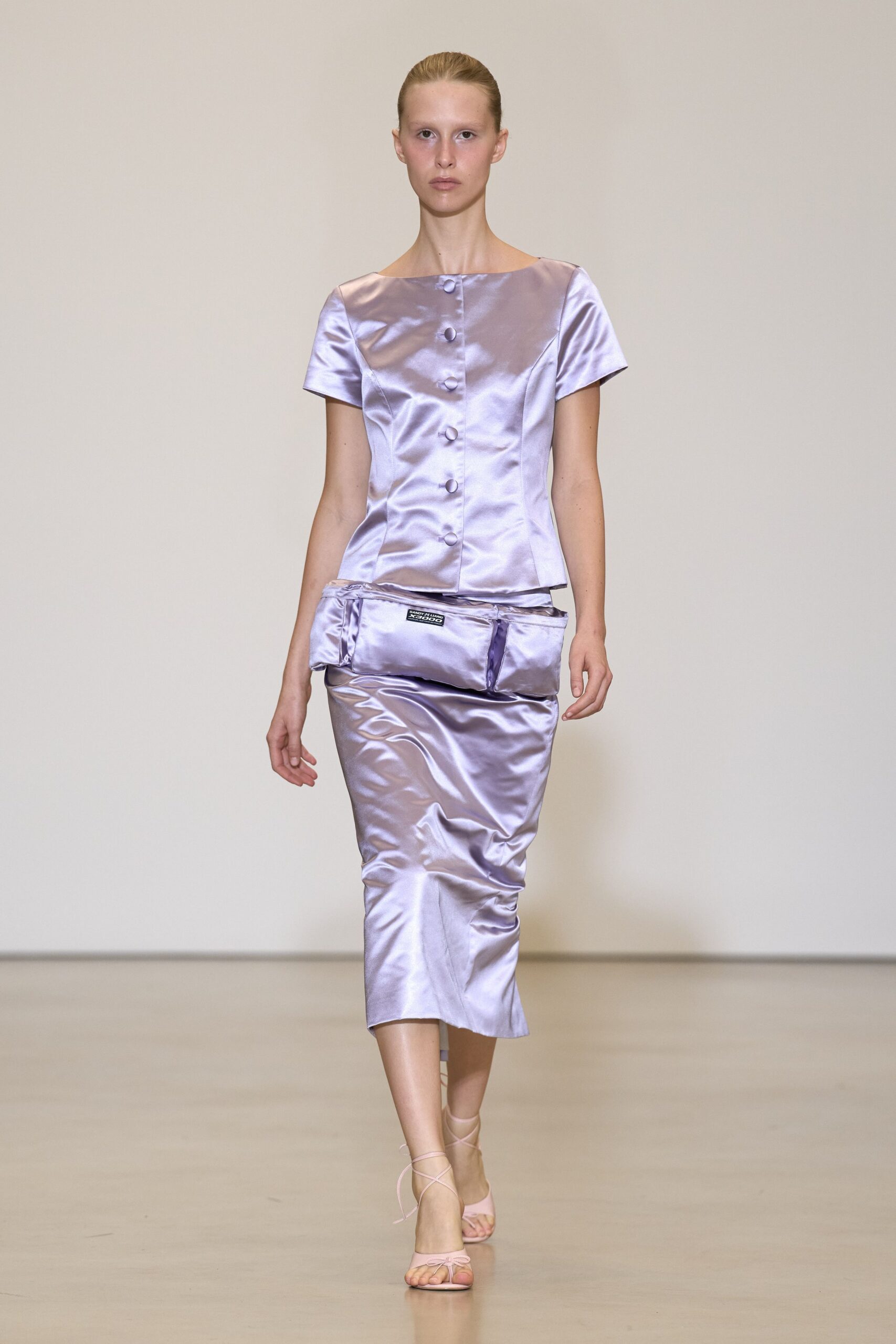 Lilac duchesse satin top with a bateau neck and covered buttons. Padded satin belt bag slung low, label patch centered. Matching satin pencil skirt, lightly ruched and kicked at the hem. Pale pink lace-up sandals. For carrying cake boxes home after a courthouse wedding.