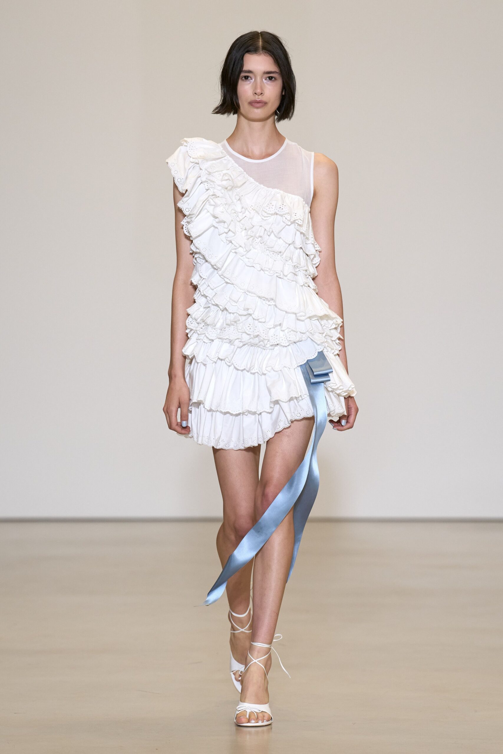 White cotton minidress with sheer one-shoulder yoke and tiered broderie ruffles. Powder-blue satin bow trailing in a long loose tail. White lace-up sandals with narrow straps crossing the instep. Walking across a hotel lawn after the wedding chairs are stacked.