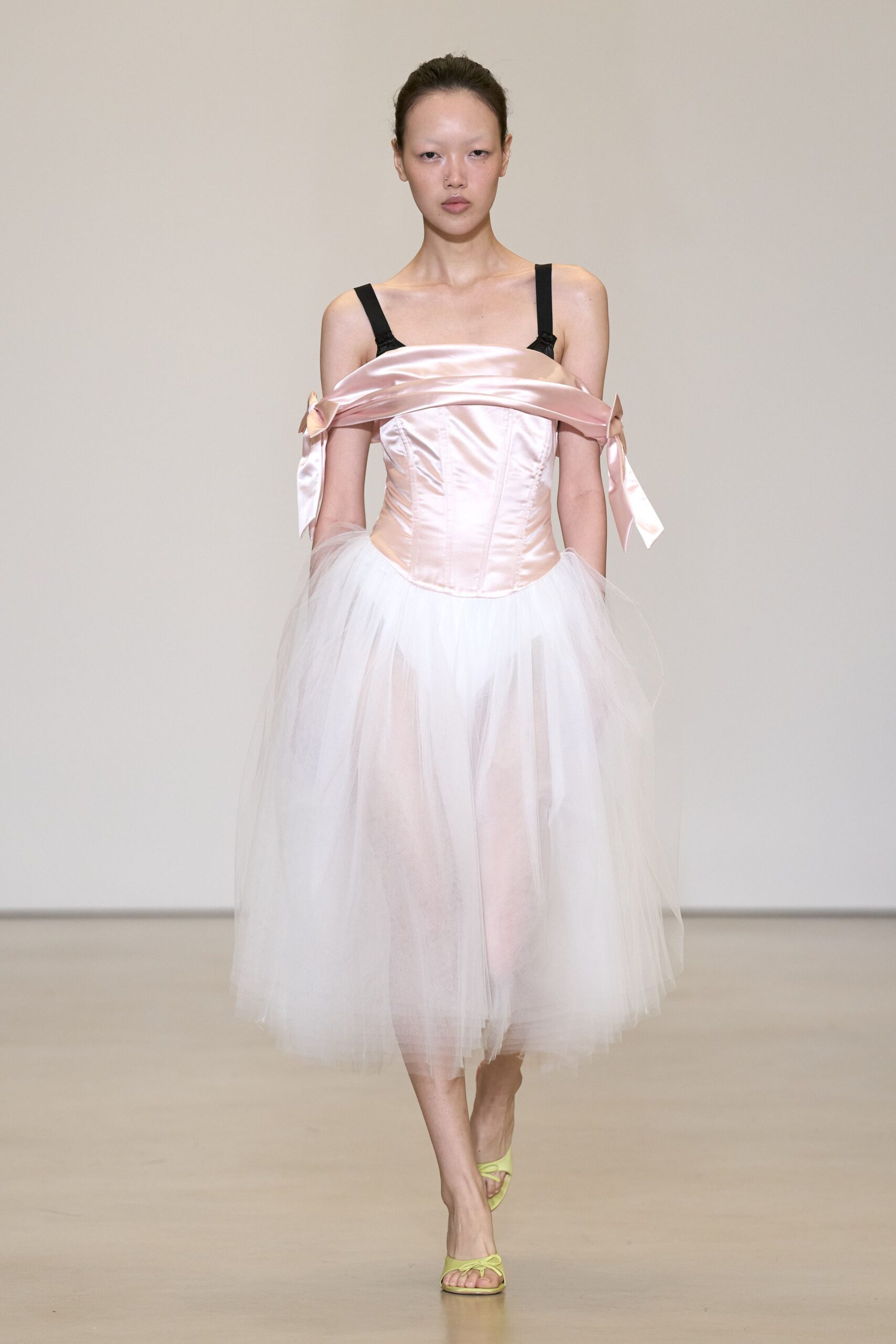 Blush satin corset dress with dropped foldover neckline and bow-tied sleeves. Black shoulder straps interrupting the pale bodice. Cloudy white tulle falling sheer over the legs. Lime satin thong sandals with a tiny bow at the toes. After the recital, still half dressed in the practice room mirror.