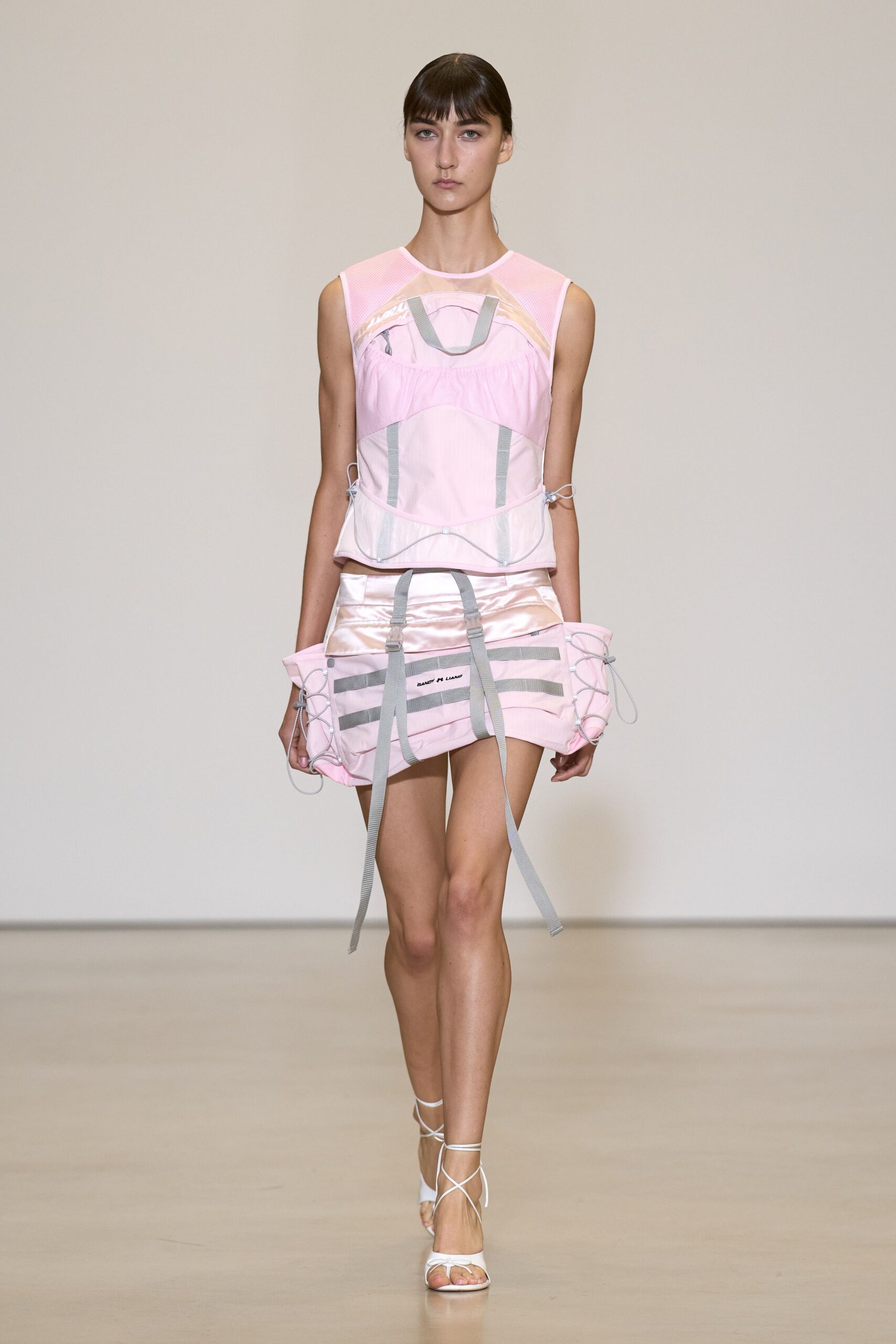 Sheer blush mesh top panelled with satin bands and grey webbing loops. Bubblegum satin mini skirt cinched wide with dangling seatbelt straps. White lace-up sandals with thin cords crossing the bare foot. Walking the marina boardwalk after buying nothing but a pink sports drink.