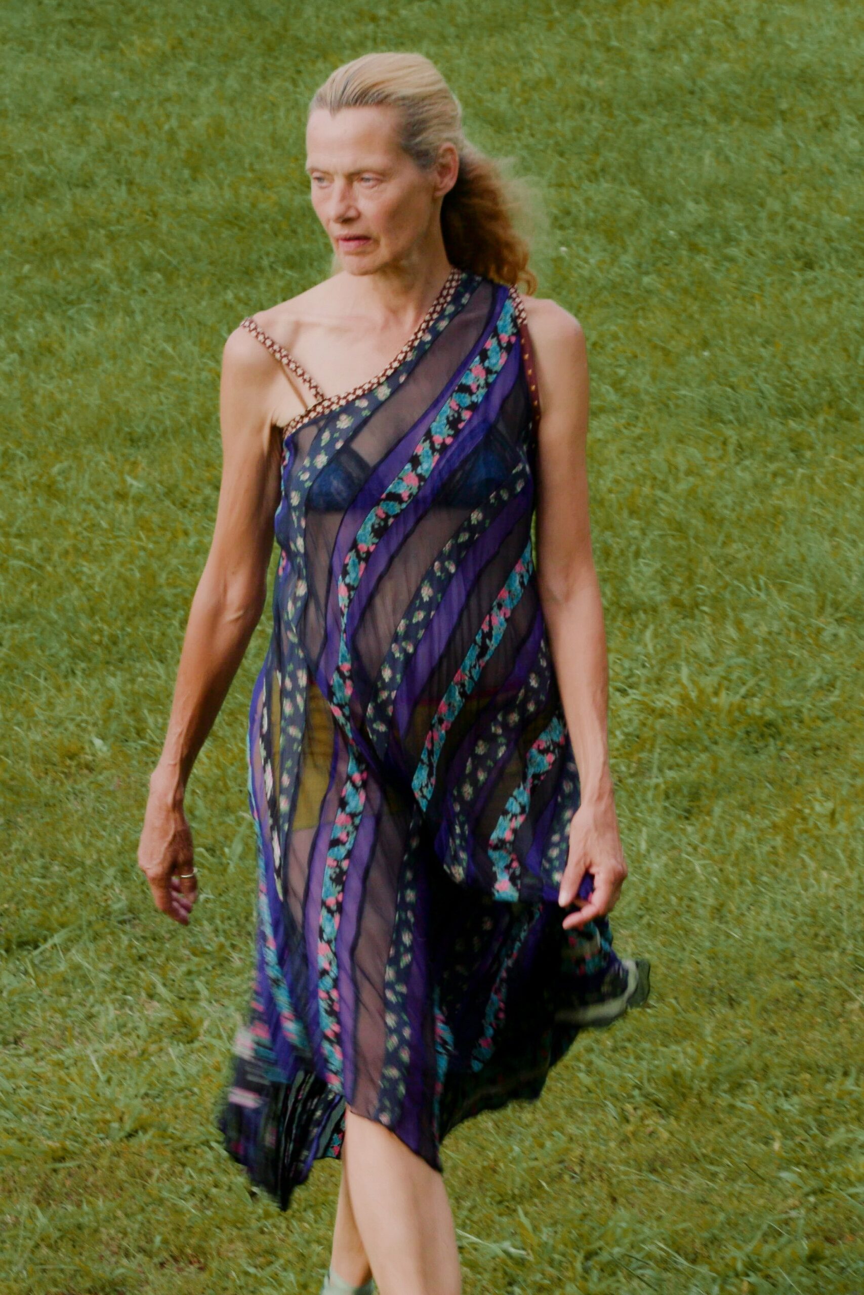 Sheer one-shoulder dress in black chiffon, pieced with violet bands and tiny floral ribbons. Crossing a wet lawn after the wedding tents have come down.