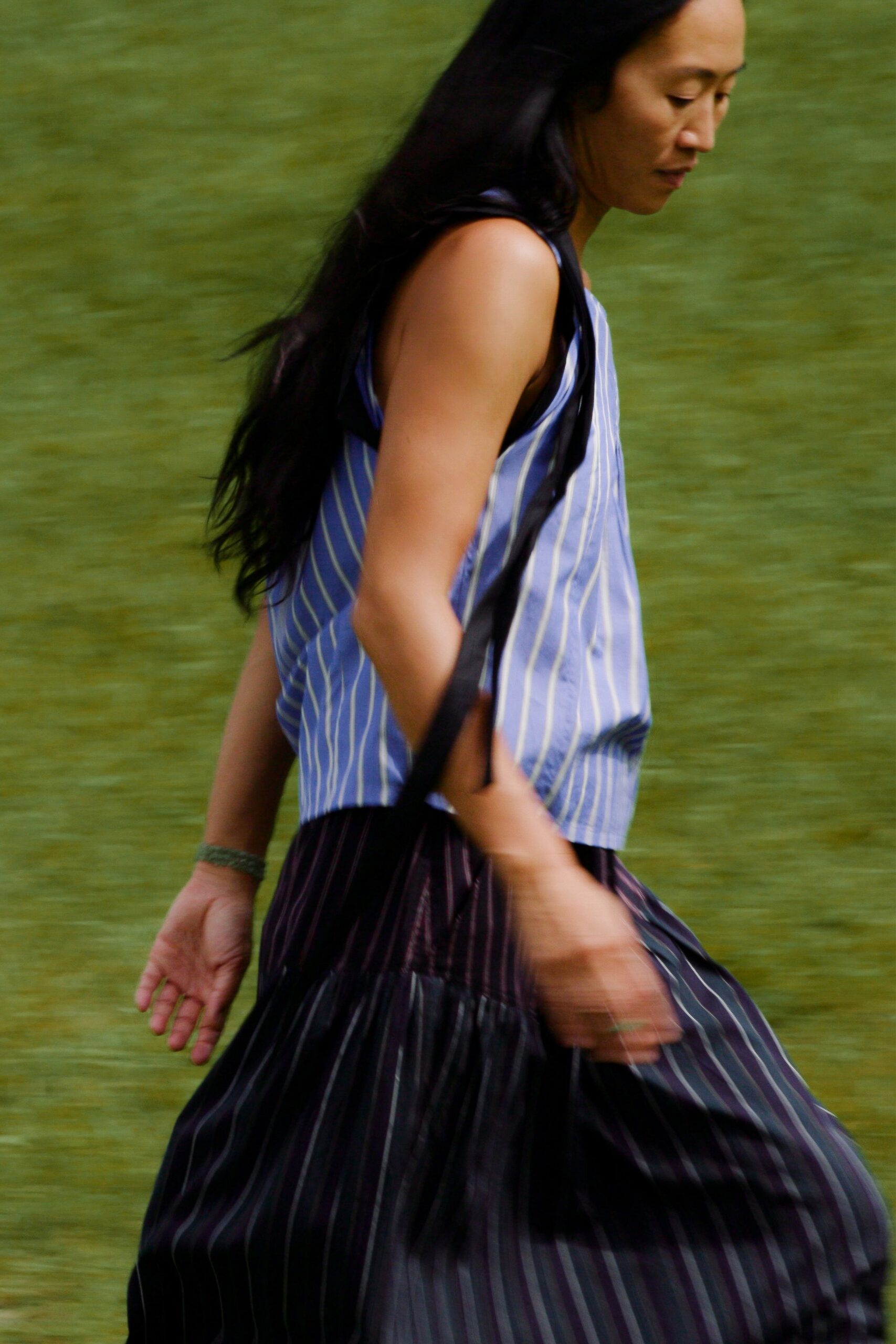 Powder-blue striped shell top with deep armholes and black binding. Black pinstriped skirt cut full, pleats loosening into motion. Moss-green band at one wrist. Walking beside a lake fast enough for the hem to keep swaying.