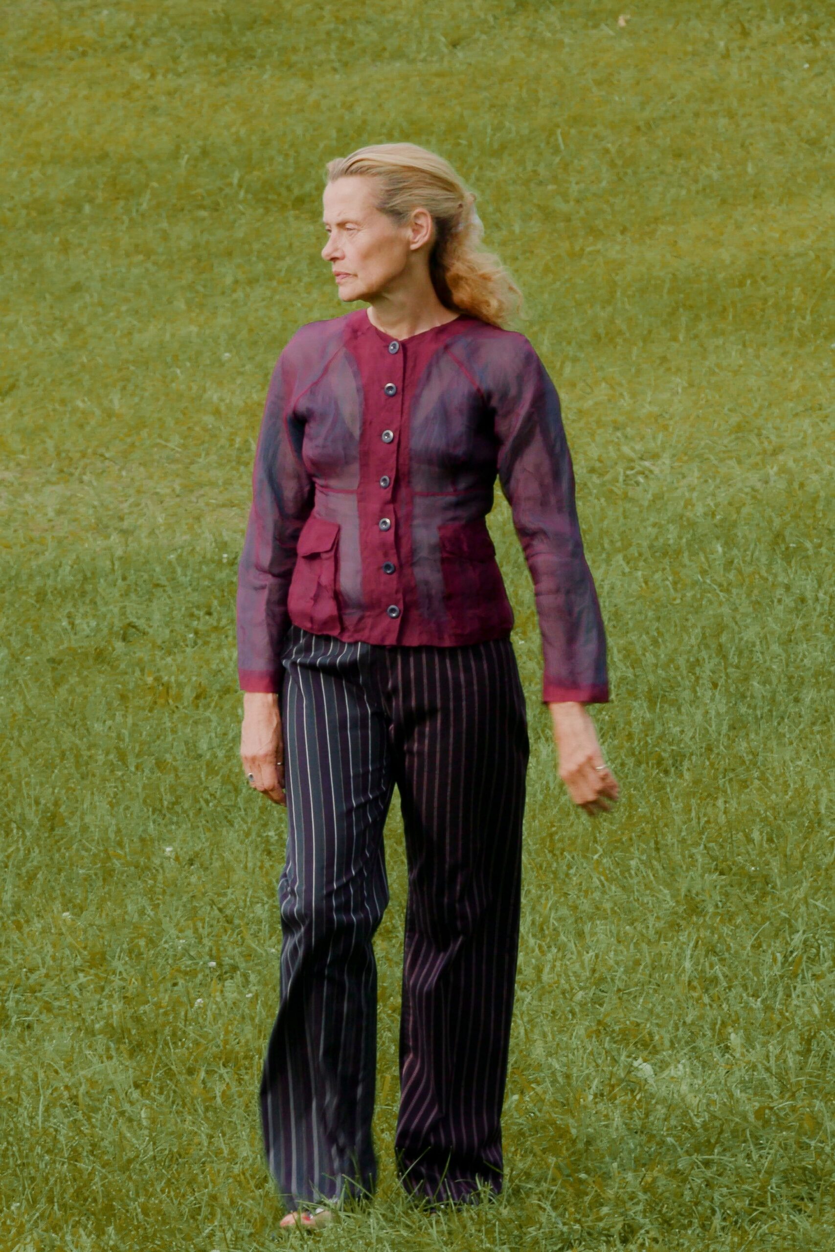 Sheer oxblood blouse with smoky vertical shading and mismatched dark buttons. Black pinstriped trousers falling straight from the hip. Flat sandals, a glimpse of red at the toe. Crossing a wet lawn after the wedding has already thinned out.