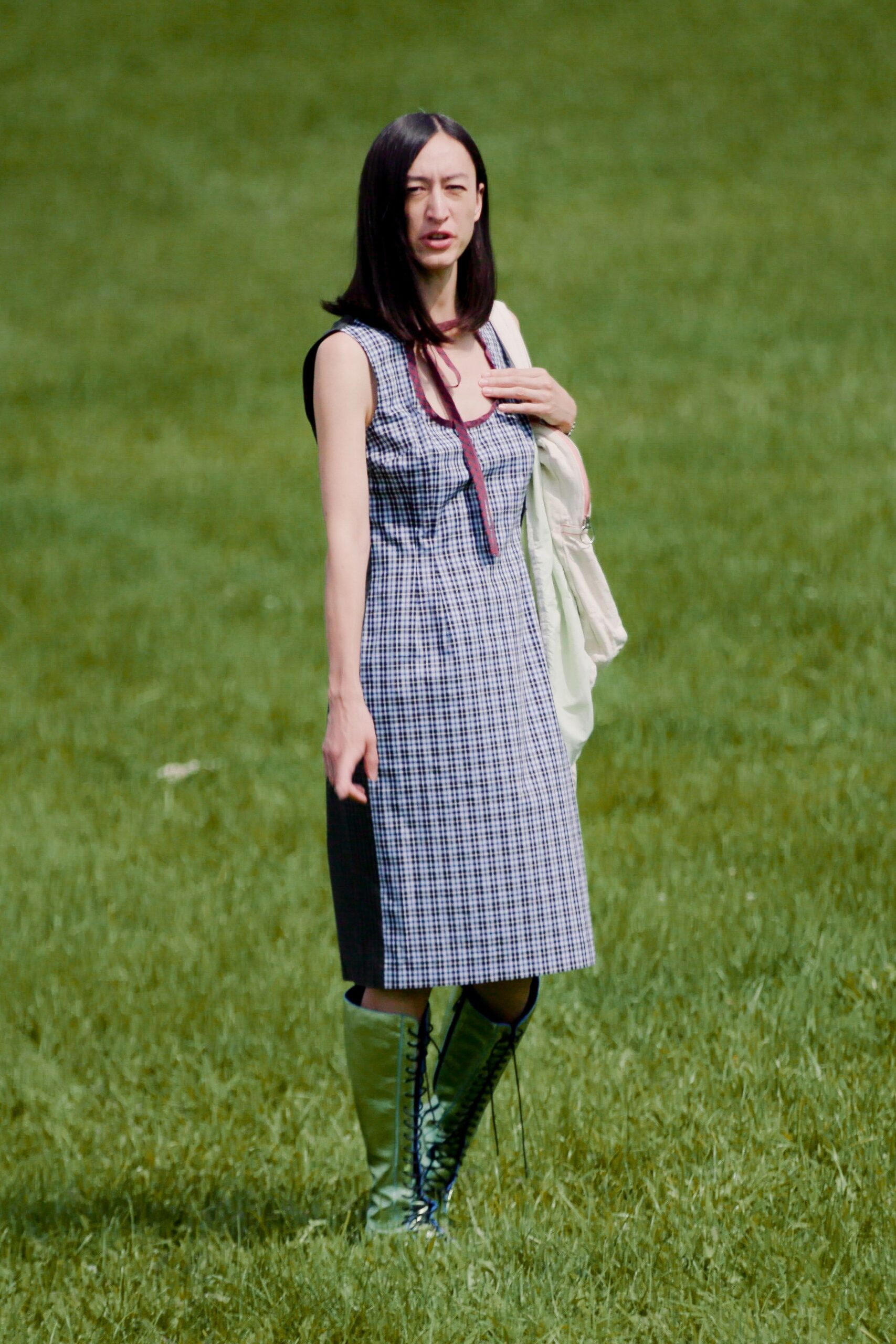 Black-and-white micro-gingham tank dress, burgundy binding trailing into a neck tie. Slouchy white shoulder bag with a ring pull catching at the side. Olive knee-high lace-up boots with glossy shafts and loose black cords. Crossing a wet meadow after lunch, when the ground keeps taking your heel.