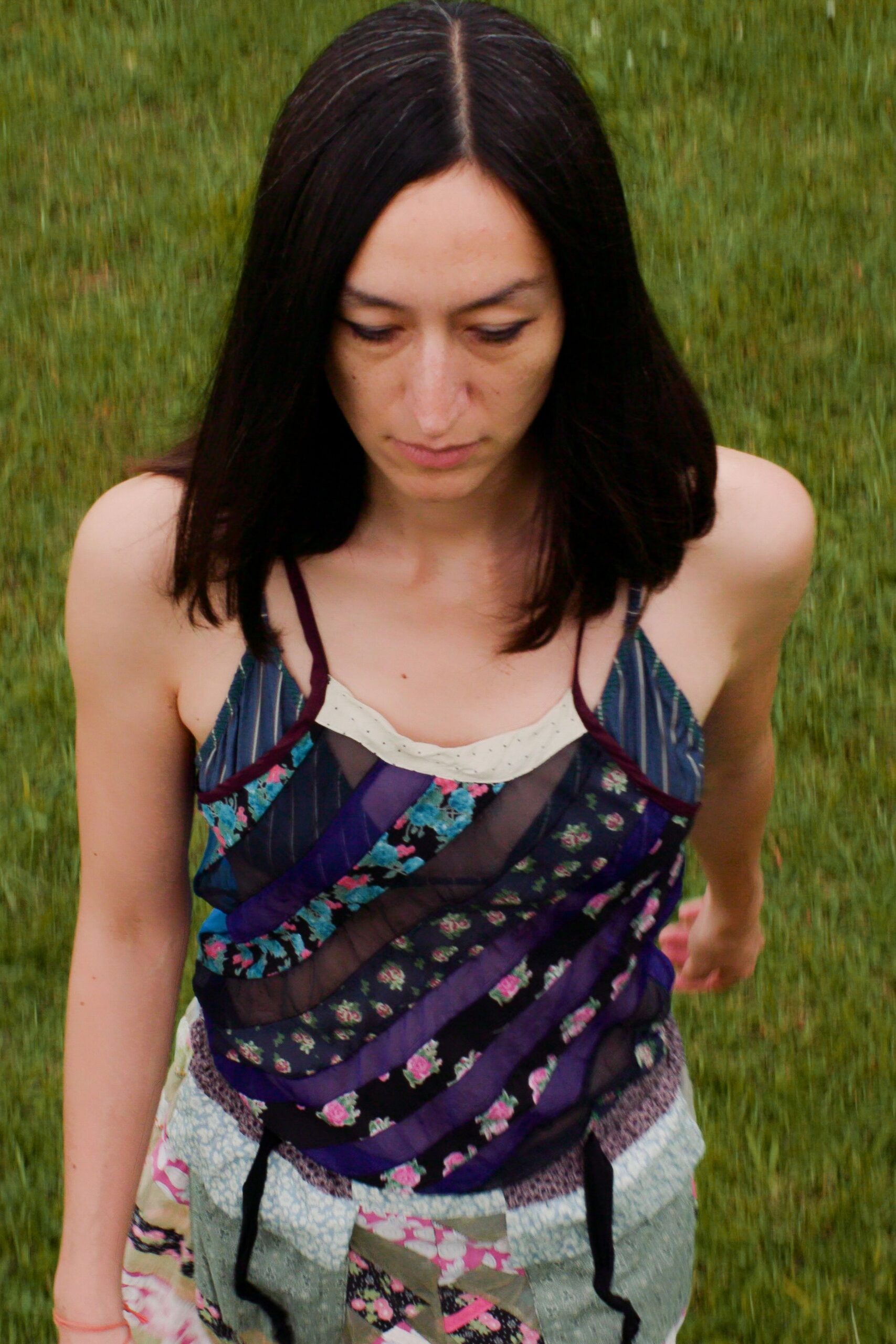 Bias-pieced camisole in mixed floral scraps with sheer black bands. Cream inset peeking at the neckline, tiny dots along the edge. Patchwork skirt in washed florals and sage panels, black ties hanging loose. Crossing a damp lawn after the picnic blankets were folded.