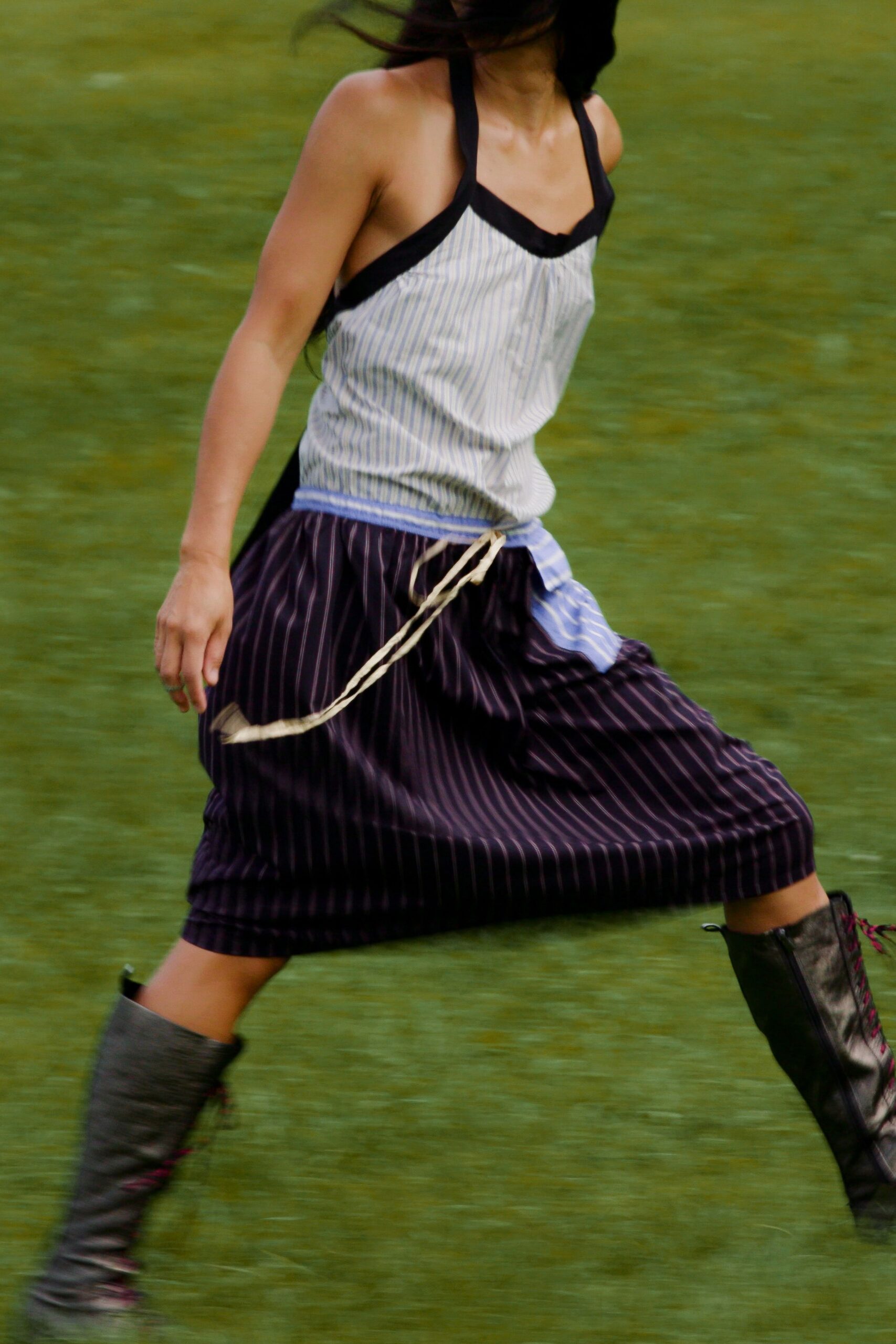 Blue ticking-stripe halter top with black binding and a ruched center seam. Dark plum pinstripe culottes, waistband patched in mismatched shirting with rope ties swinging loose. Black knee boots laced in magenta cord. Running across wet grass before the picnic blanket blows away.