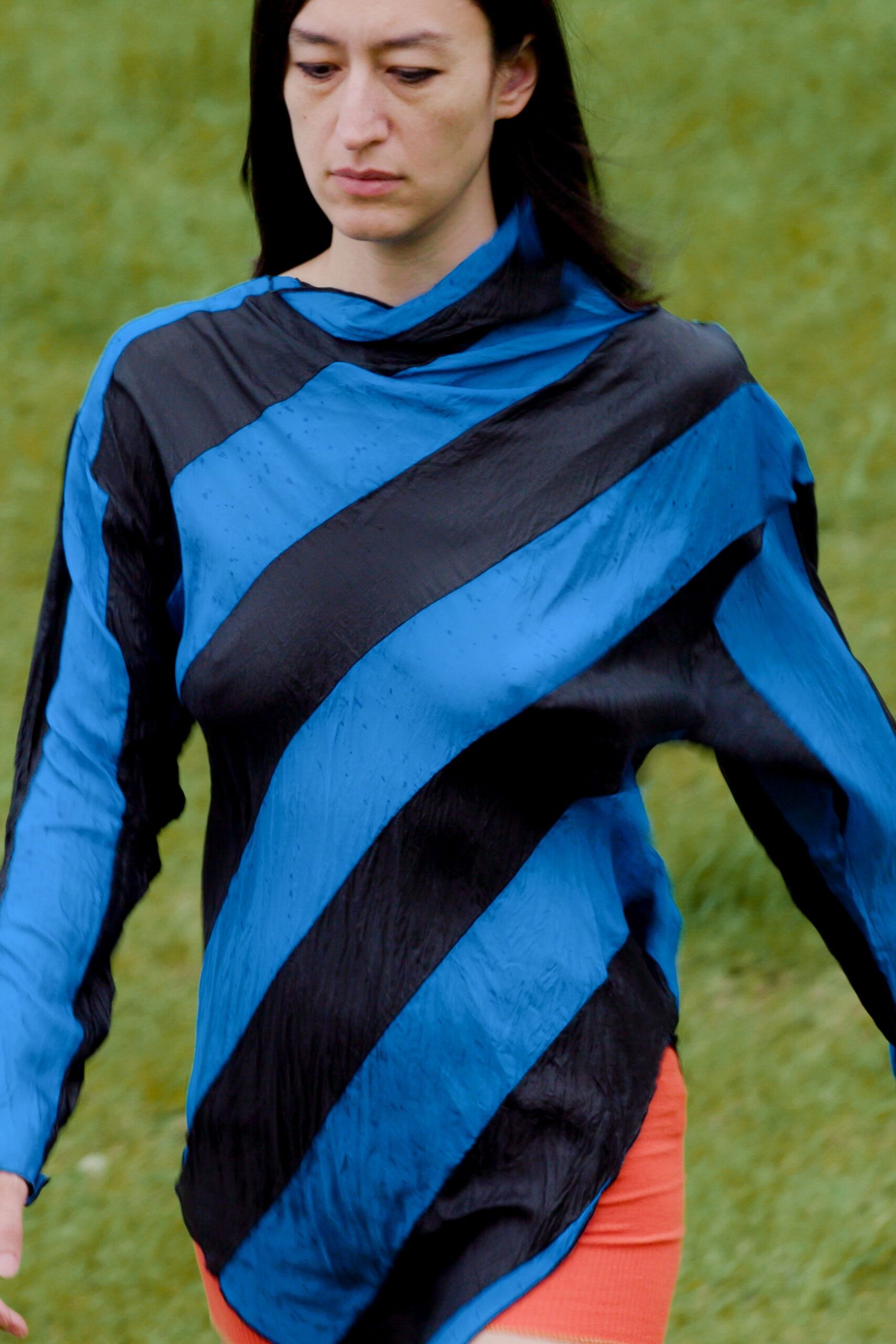 Crinkled nylon top in cobalt and black diagonal bands, neck softly twisted. Ribbed coral knit hem peeking below like a second layer. Crossing wet grass after the lake wind turned cold.