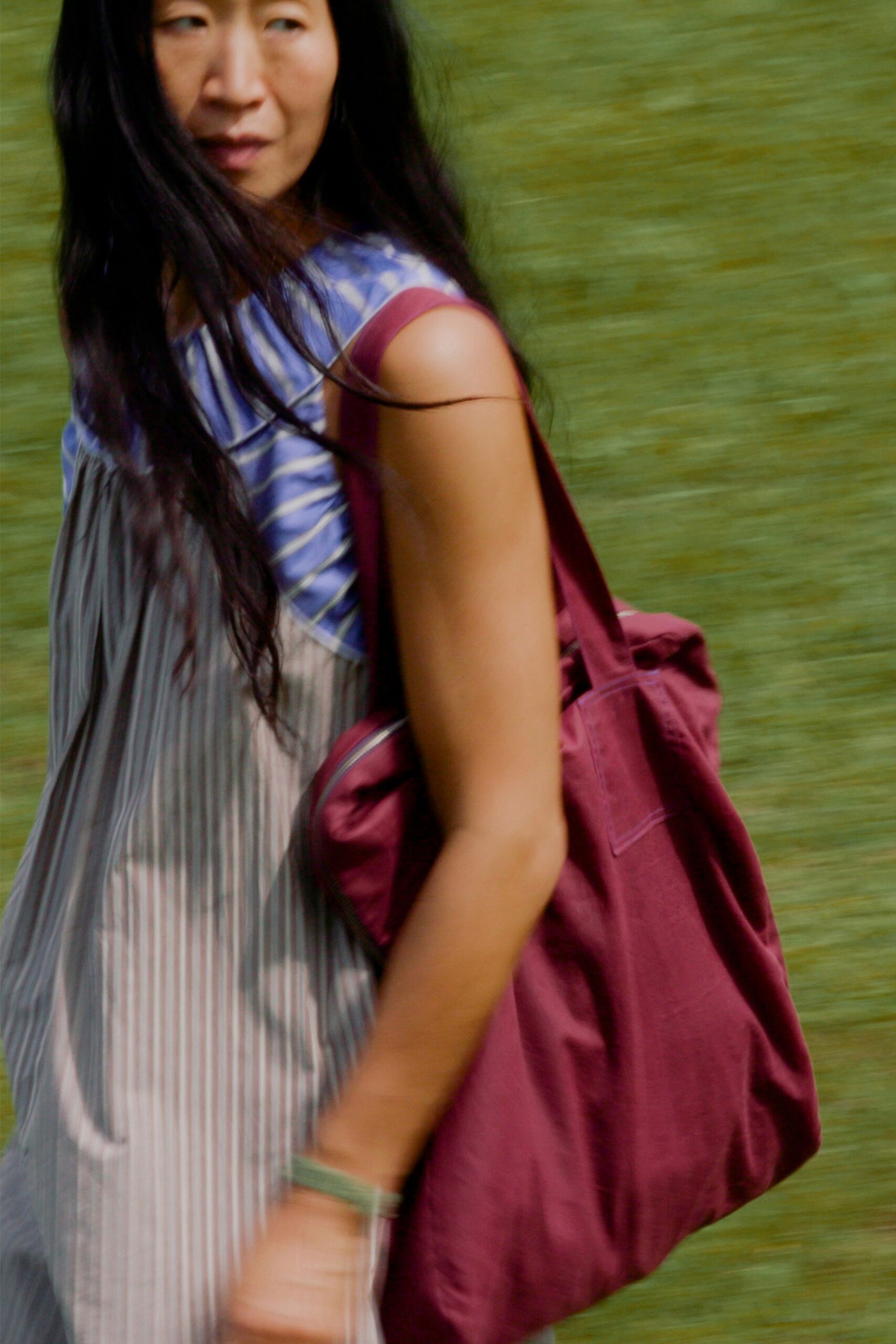 Blue-and-white marbled bodice over a sheer grey pleated column. Burgundy canvas tote with a broad strap and boxed pocket. Pale green bangle, translucent and thick at the wrist. Crossing a park footpath while the picnic blanket is still being shaken out.