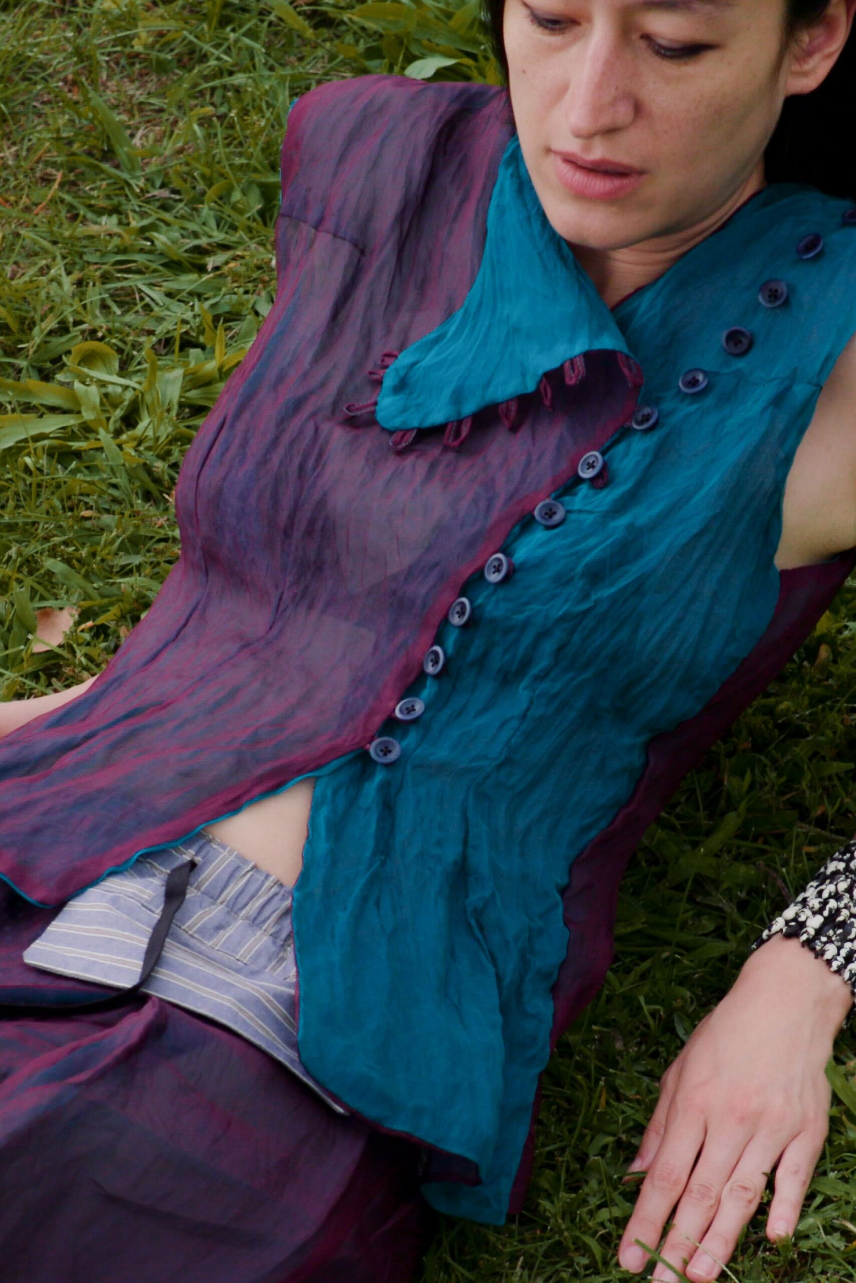 Sleeveless crinkled dress in teal and plum, double-breasted with a floppy collar. Pale striped waistband peeking out, tied with a narrow charcoal ribbon. Fallen into the grass after the picnic plates were cleared.