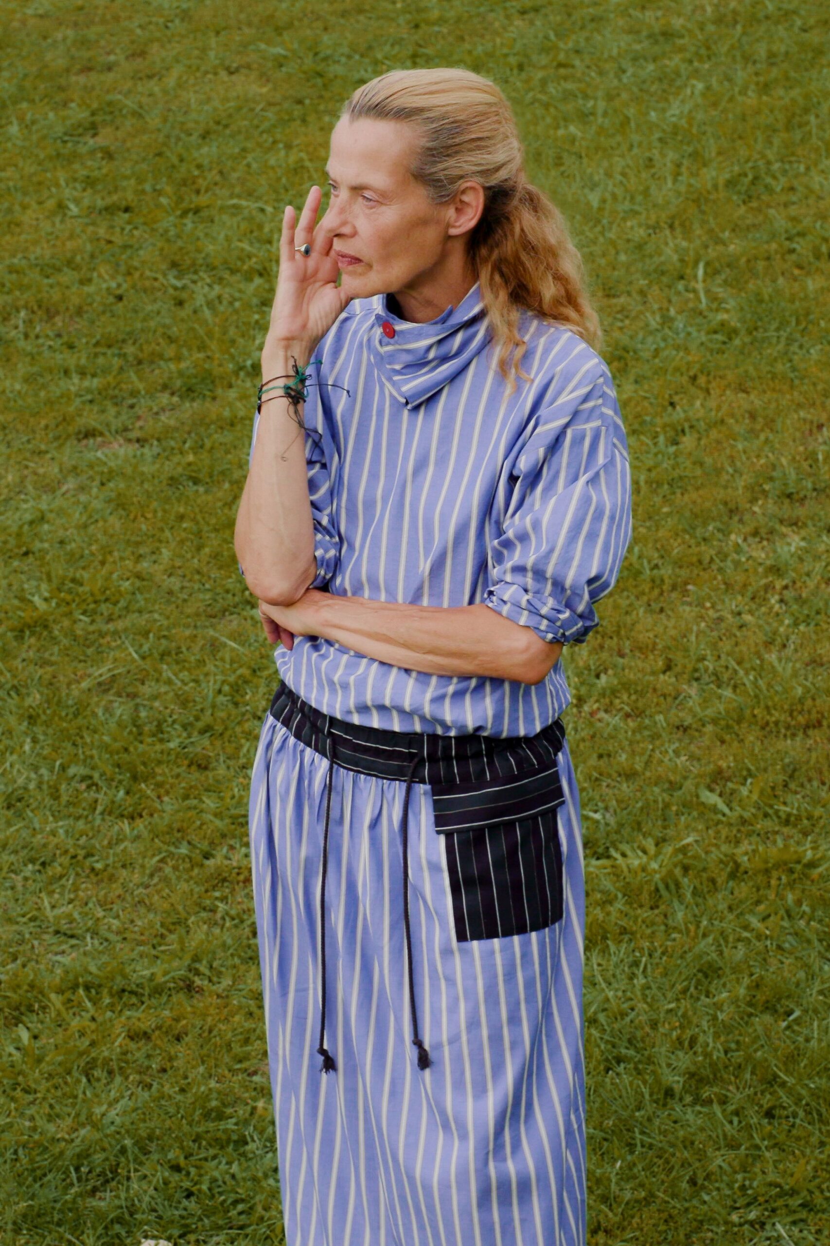 Periwinkle striped top with a loose funnel neck fastened by one red button. Matching gathered skirt cinched with a black pinstripe wrap panel and cord ties. Thin black cords knotted with a small green plastic spider. Crossing a damp lawn after the outdoor lecture has already started.