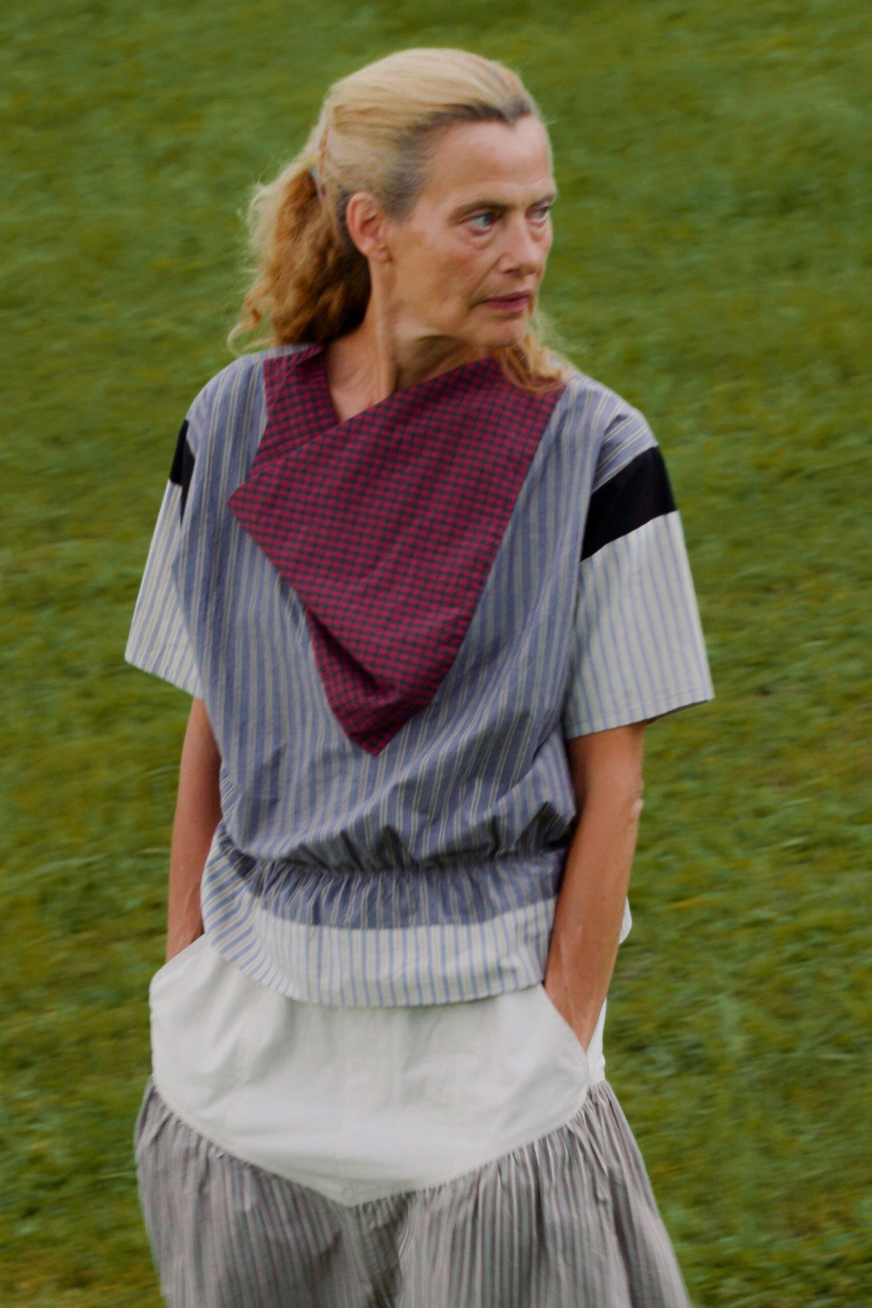 Blue ticking-stripe top with dropped shoulders and a gathered blouson waist. Burgundy micro-check kerchief folded into a sharp bib over the chest. Pale striped skirt with a clouded white panel and soft pleats below. Crossing a damp lawn after lunch, still carrying the picnic blanket.