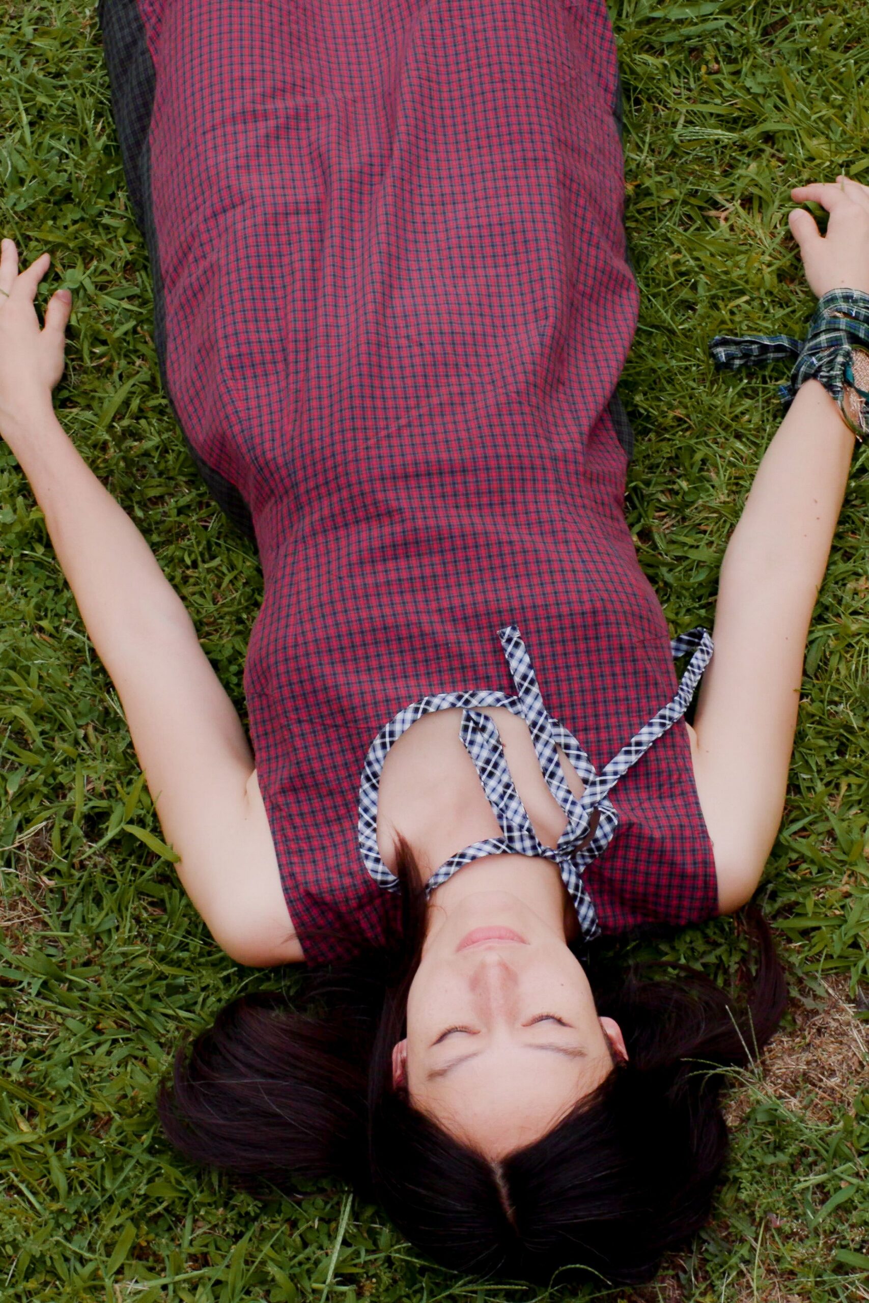Sleeveless column dress in crimson-and-navy microcheck, cut with a deep keyhole neckline. Blue gingham straps looped and knotted loosely across the throat. Matching gingham strips tied at both wrists, one end jutting out. Afternoon grass still damp after the picnic blankets were folded away.
