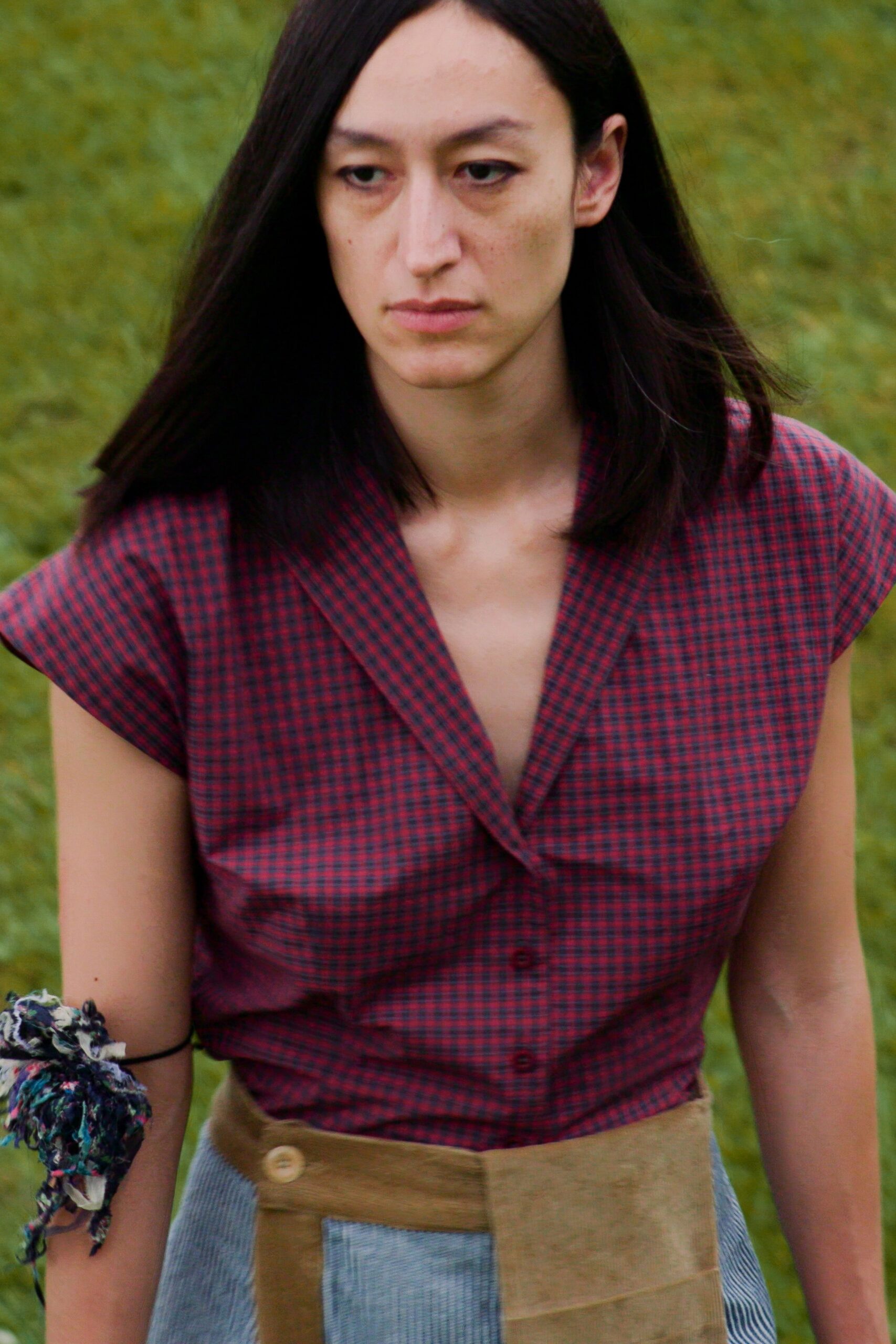 Red-and-navy microcheck blouse with cap sleeves and a low revere collar. Patchwork skirt in tan suede and pale blue pinstripe panels, buttoned at one side. Shredded floral fabric knotted tight around the upper arm. Walking across wet grass after the picnic blankets have been folded.