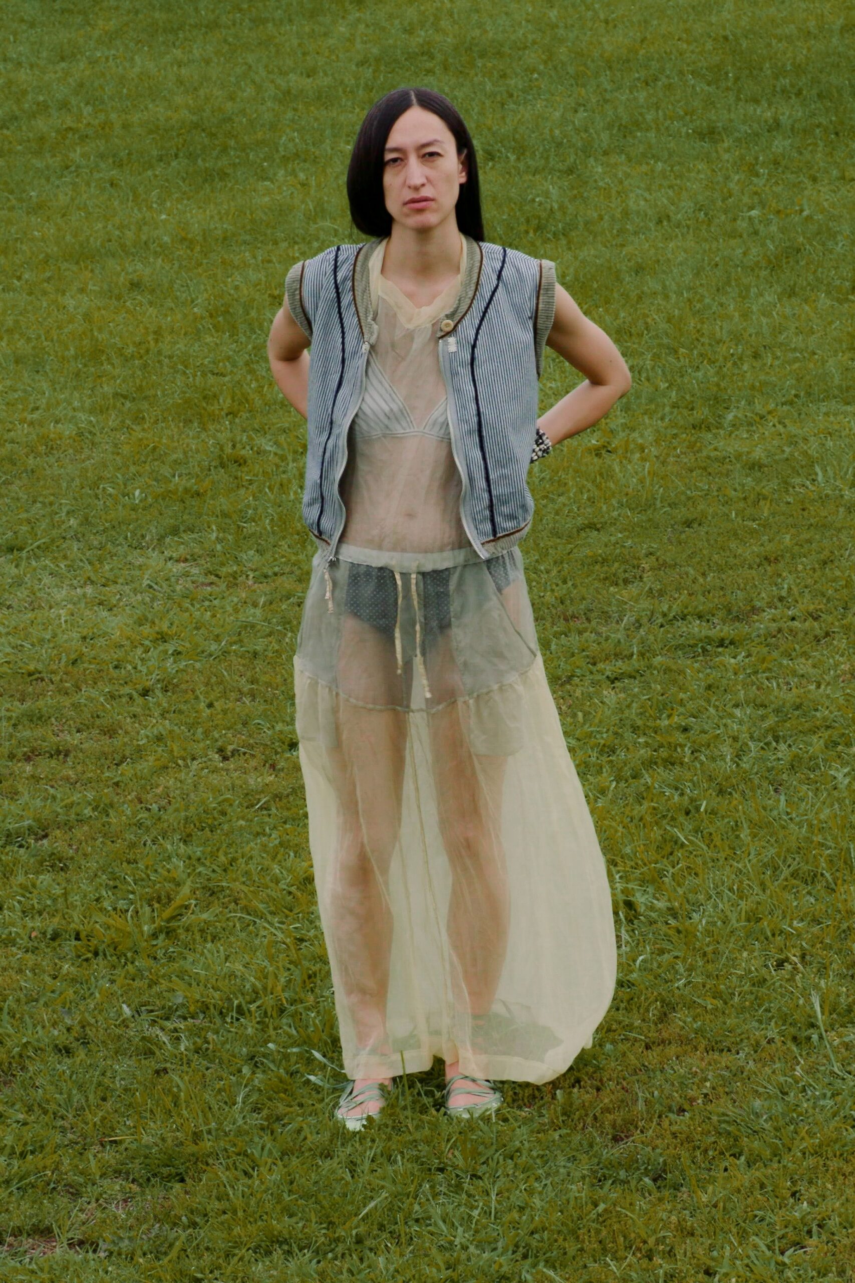 Pale blue pinstriped vest with black piping and a rope-tassel zip. Sheer butter chiffon dress, gathered under a soft bralette seam. Charcoal dotted briefs visible through the skirt. Mint lace-up flats wrapping low across the foot. Chunky silver beaded bracelet. Crossing wet grass after swimming, towel still on the bank.