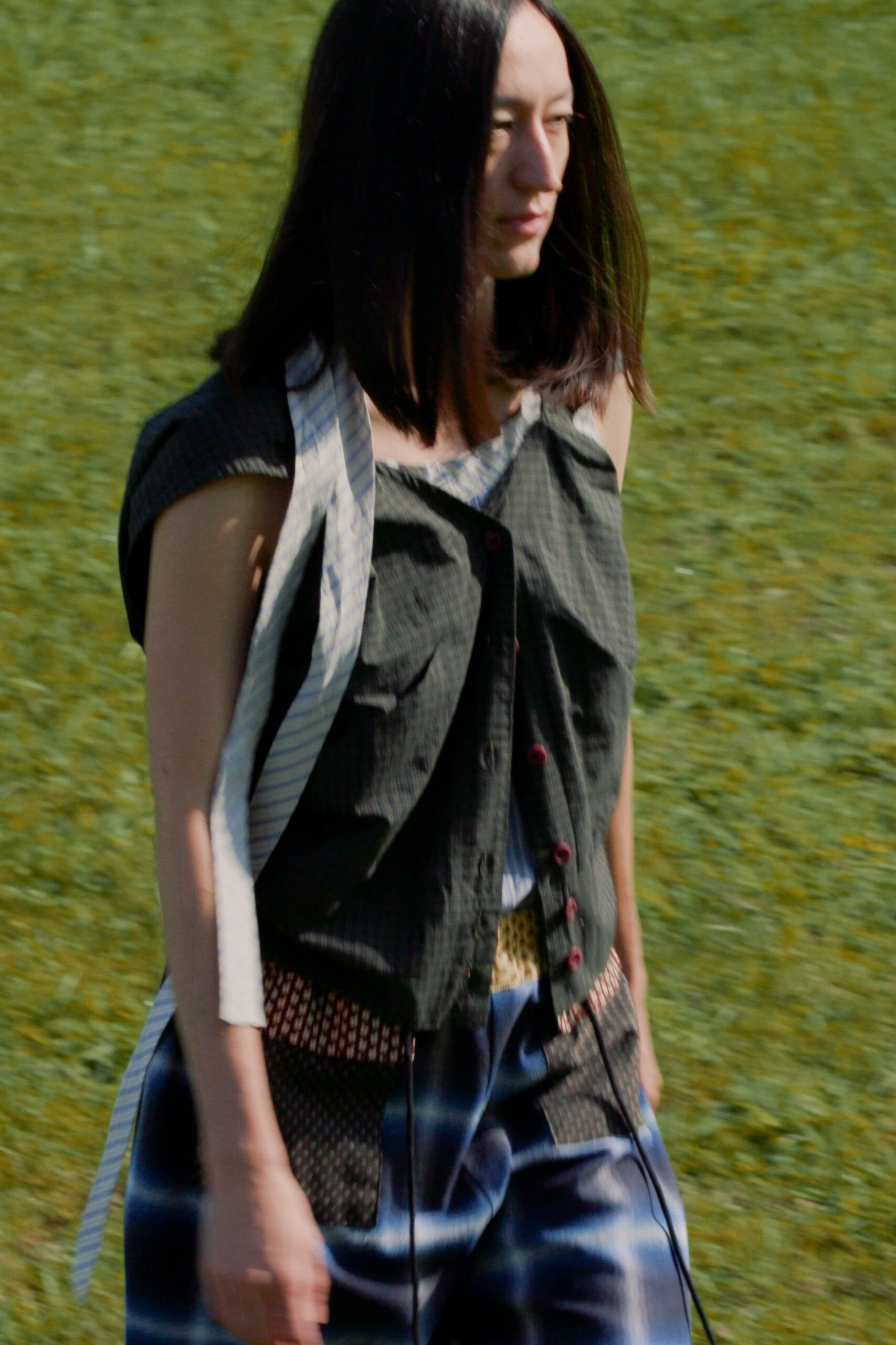 Charcoal pinstriped sleeveless top, crookedly buttoned with wine-red buttons. Pale blue striped shirting panel hanging loose from one shoulder. Blue satin patchwork skirt with mesh inset pockets and a dangling cord. Crossing a sunburnt field after lunch, shoes damp from the grass.