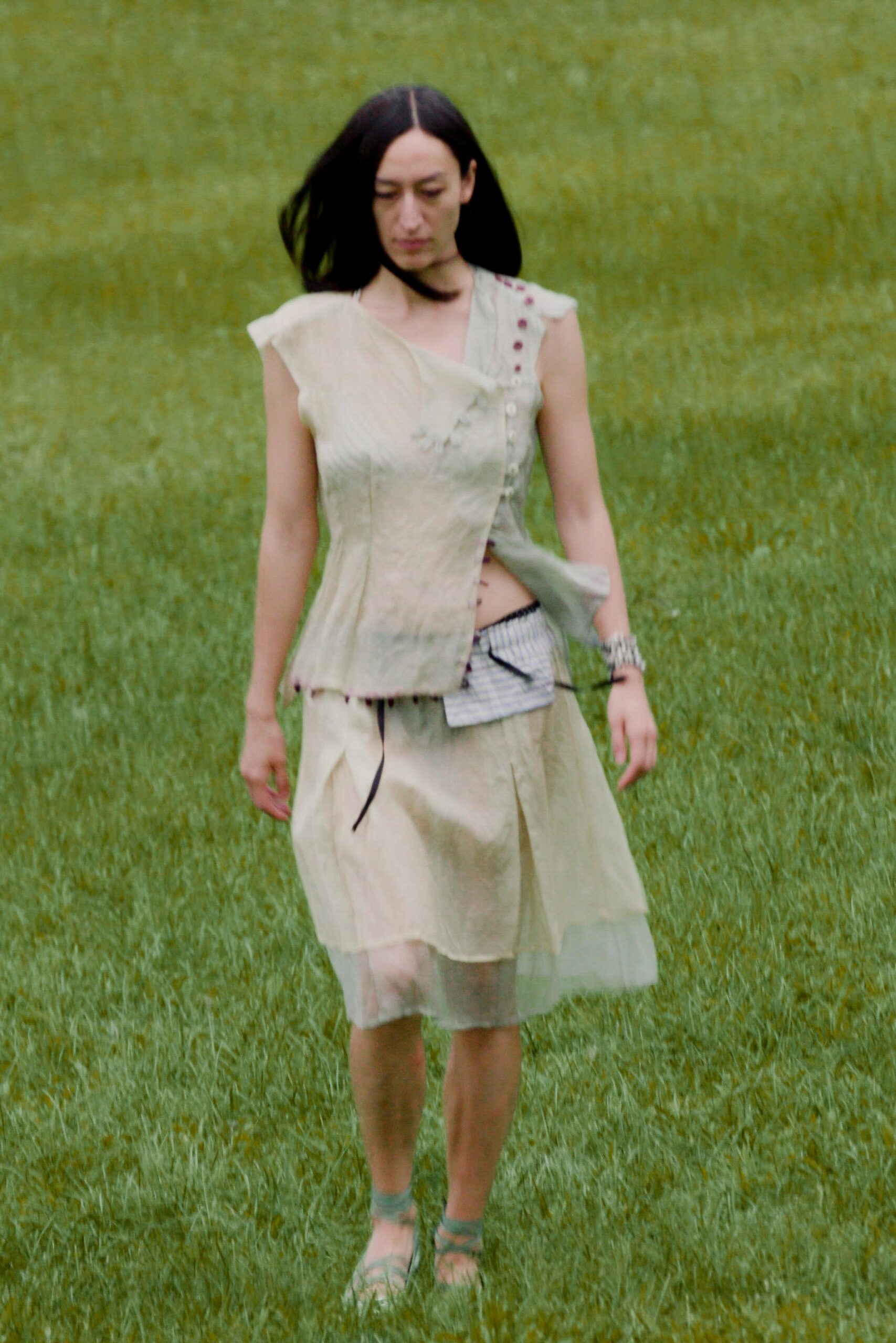 Pale sage crinkled top, skewed closure dotted with burgundy knot buttons. White pinstriped pouch tied on with black cords, one end whipping loose. Washed ivory skirt with a sheer tulle underlayer showing below. Stacked silver bangles. Seafoam wrap sandals. Crossing a wet field after the garden ceremony already blew sideways.