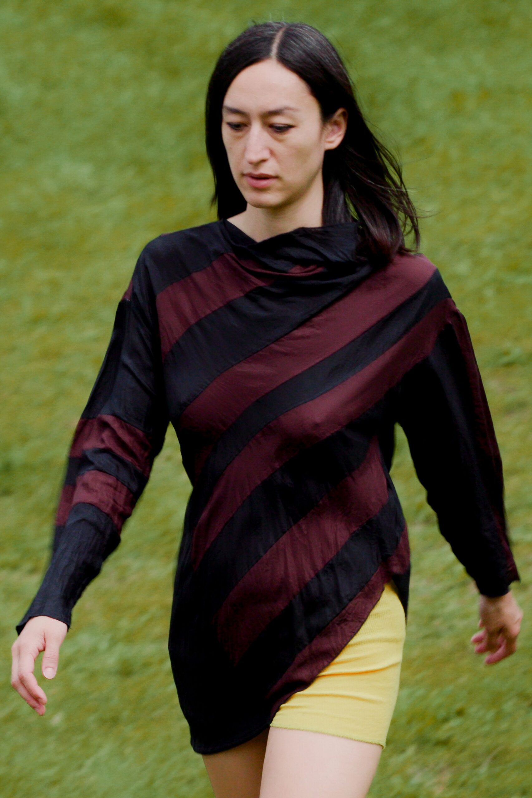 Black tunic in crinkled nylon, cut with oxblood diagonal bands and a twisted funnel neck. Lemon bike shorts in fine rib knit, showing under the uneven hem. Crossing a wet field after the fair tents have come down.