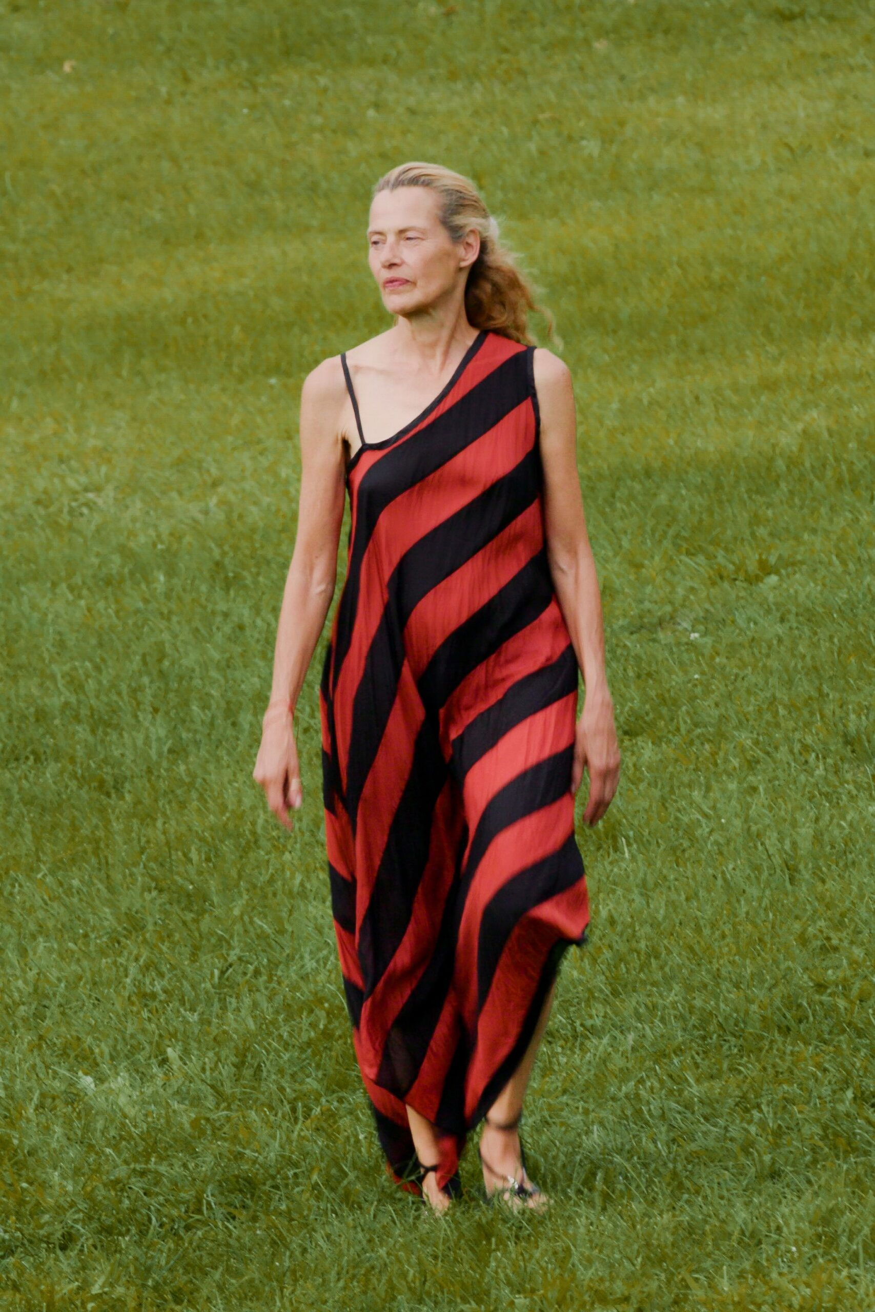 Red and black bias-striped dress in crinkled fabric, cut on a slant neckline. Black tie sandals, cords crossing low over the toes. Crossing a wet lawn after the wedding moved outside again.