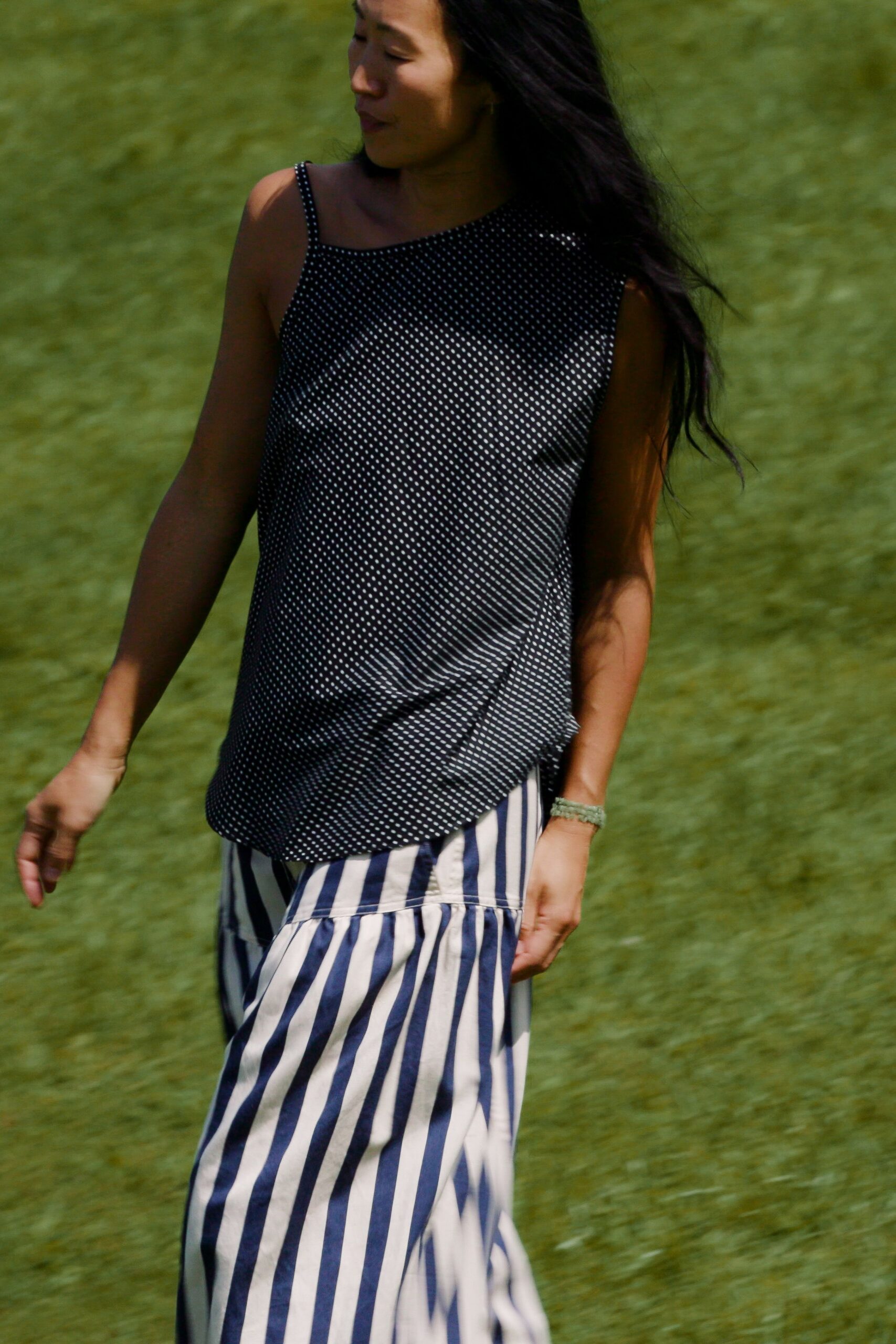 Black satin camisole peppered with tiny white dots, hem curving over the hip. Cream and navy striped skirt with a dropped seam flounce. Pale green beaded bracelet. Walking across cut grass after lunch, shoes in one hand somewhere off frame.
