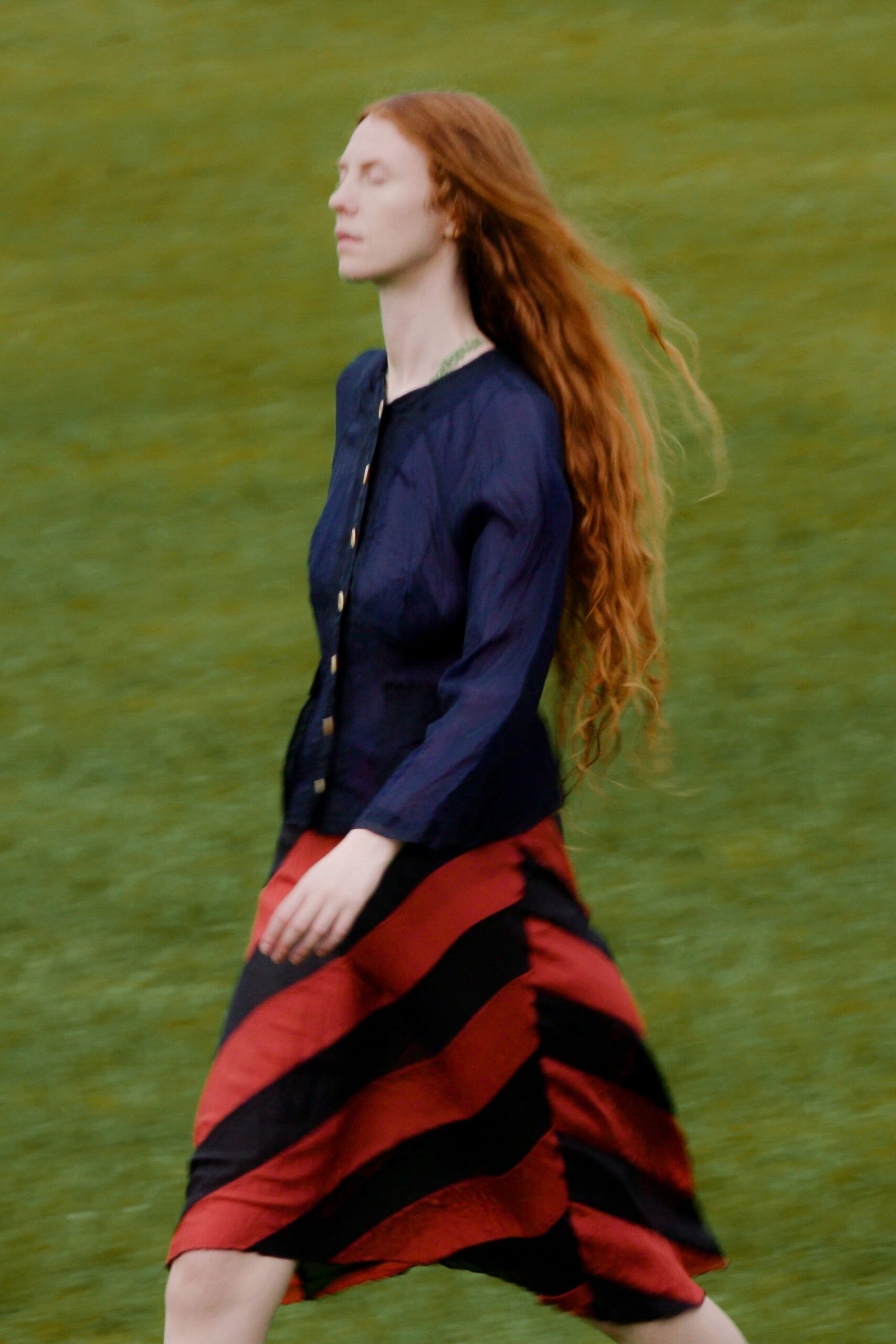 Inky crinkled blouse with a round neck and small gold buttons. Bias-cut midi skirt in rust and black diagonal bands, hem kicking outward. Crossing a wet lawn after the music has already started.