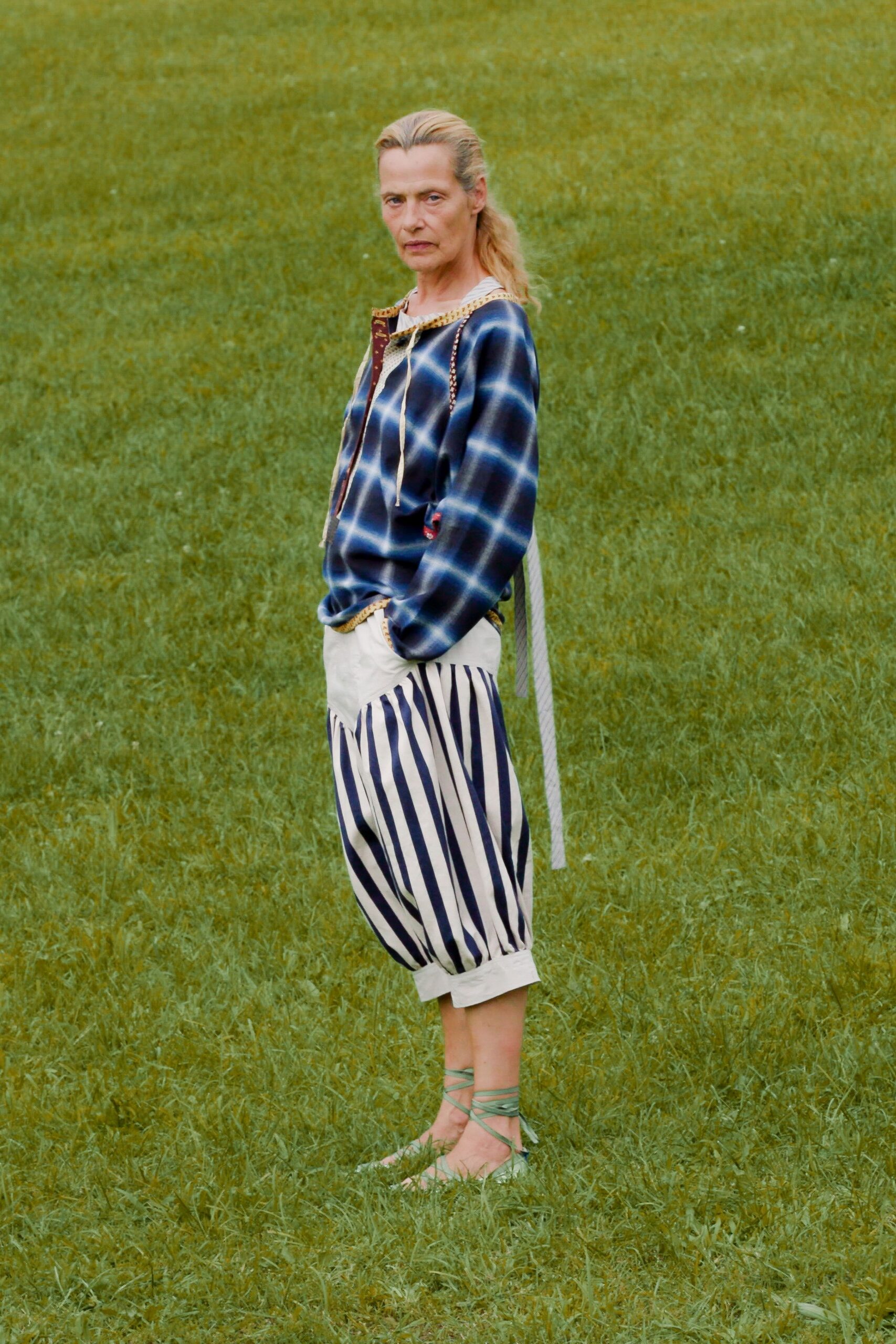 Indigo plaid hooded blouse with blanket-stitch trim and long dangling ties. Cream balloon shorts paneled with navy deck stripes, cuffed below the knee. Seafoam wraparound flats, laces crossed up the bare ankle. Walking the field before rain with the picnic still unpacked.