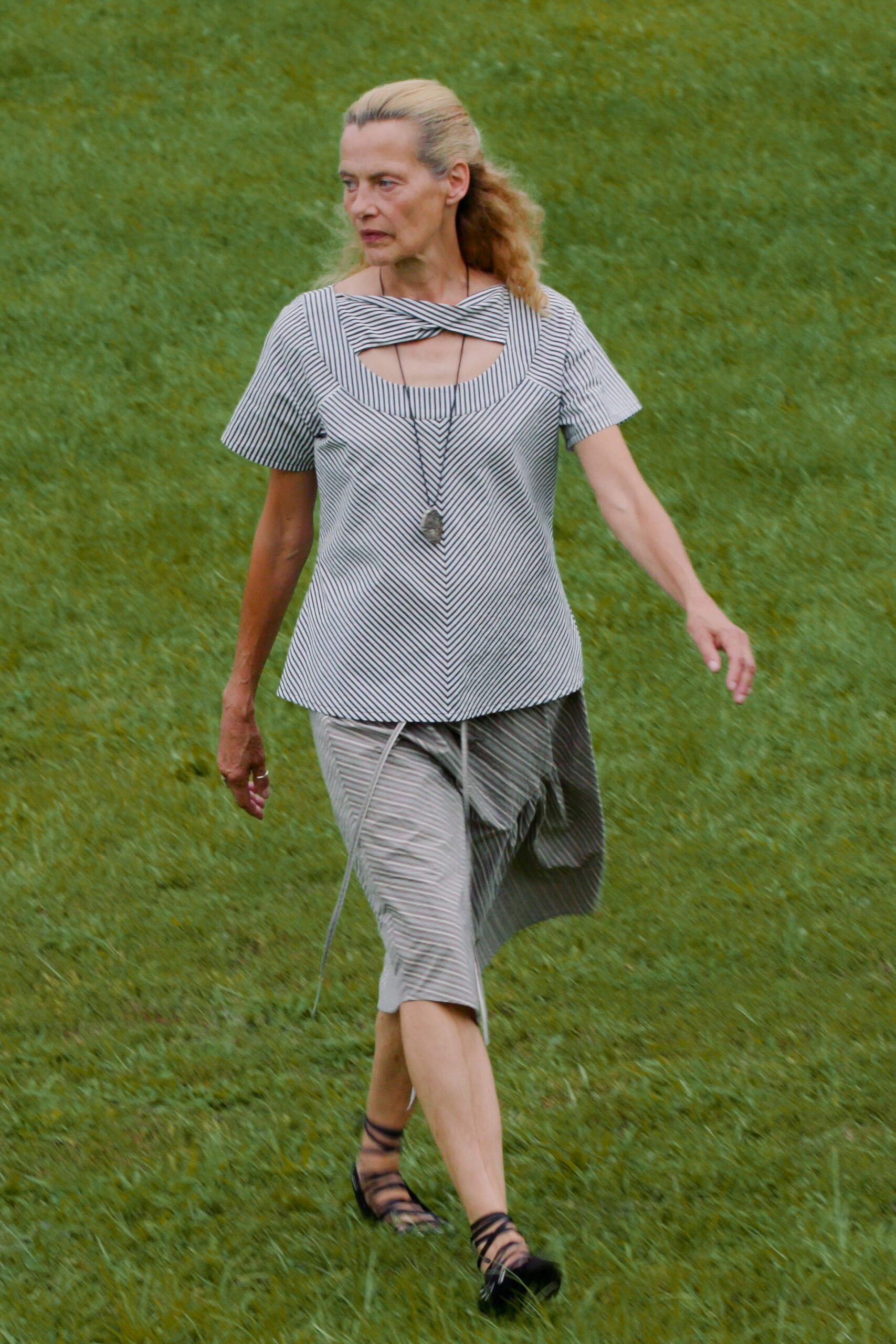 Black-and-white striped top with a twisted bandshell neckline cutout. Long cord necklace ending in a rough gray stone. Bias-cut striped skirt, one draped panel kicked loose at the thigh. Black lace-up sandals wrapping the ankle. Crossing wet grass after lunch at a sculpture park.