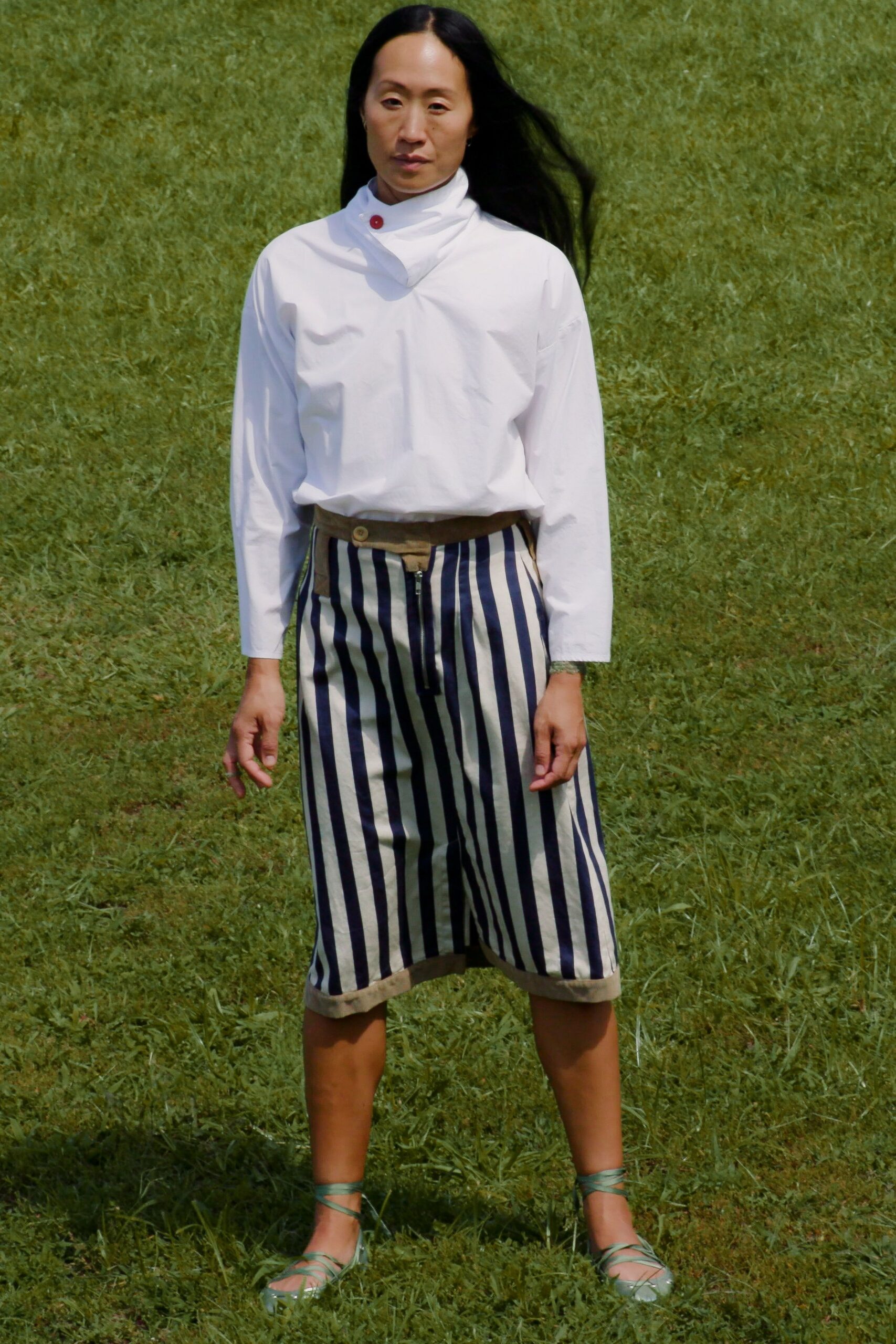 White cotton blouse with a sculpted funnel neck, one red collar button. Cream culottes in wide navy vertical stripes, raw khaki waistband and zip fly. Pale sage wrap sandals laced thin around the ankles. Crossing a wet lawn after lunch, still wearing the picnic tablecloth.
