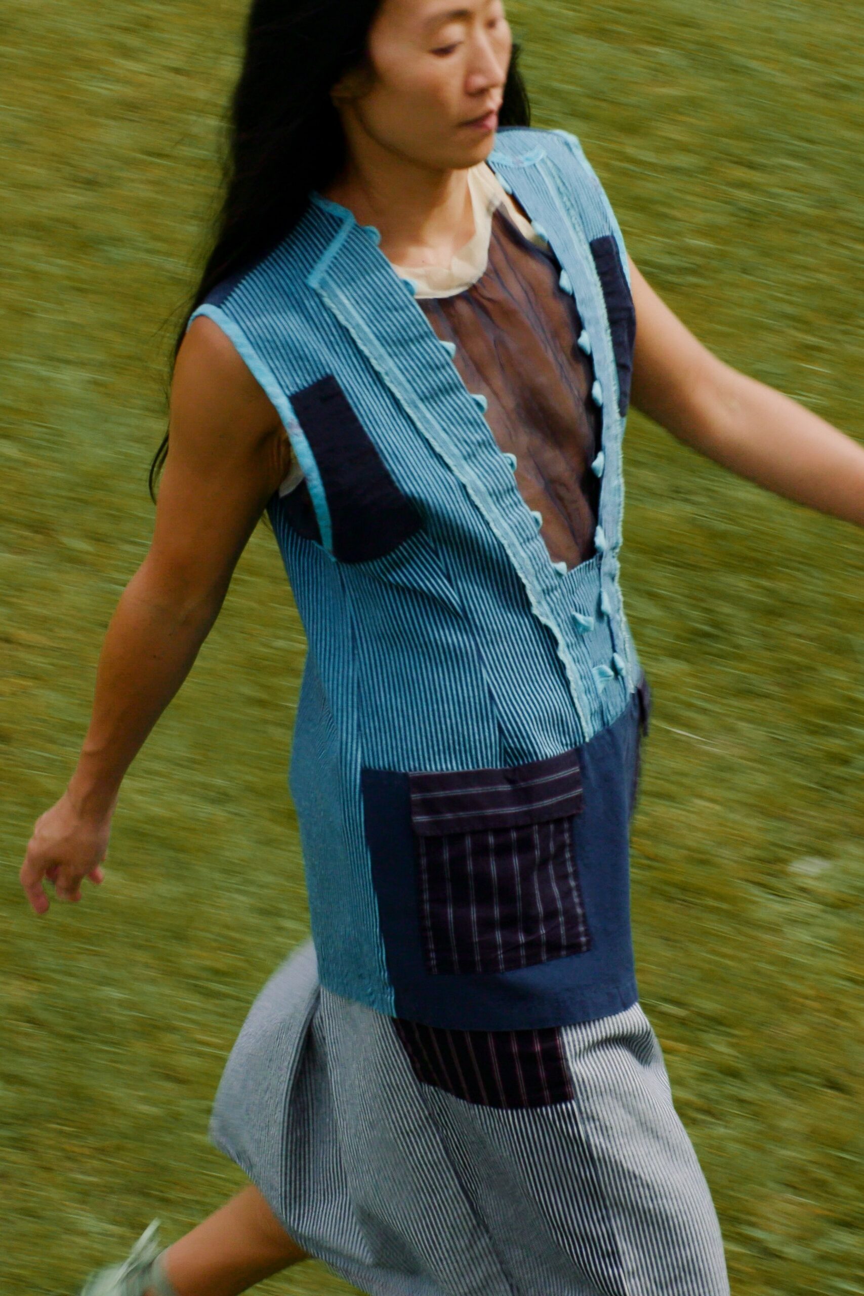 Powder-blue pinstriped patchwork dress with raw ruffled placket and bobble buttons. Sheer brown organza panel at the chest, framed by a milky collar. Mismatched navy plaid pockets, one flap sitting slightly askew. Pale mint straps, blurred mid-run. Crossing a wet lawn before the picnic blankets are claimed.