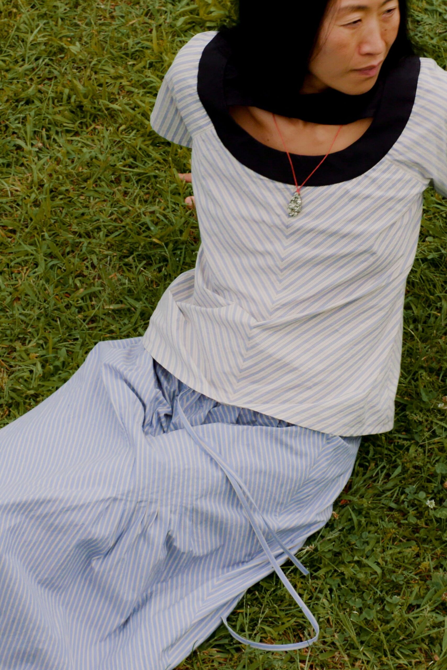 Pale blue striped cotton top with a broad black scooped collar. Black scarf looped close at the neck. Small silver pendant on a red cord. Matching striped wrap skirt with long self-ties trailing in the grass. Afternoon picnic after rain, still too damp to sit comfortably.