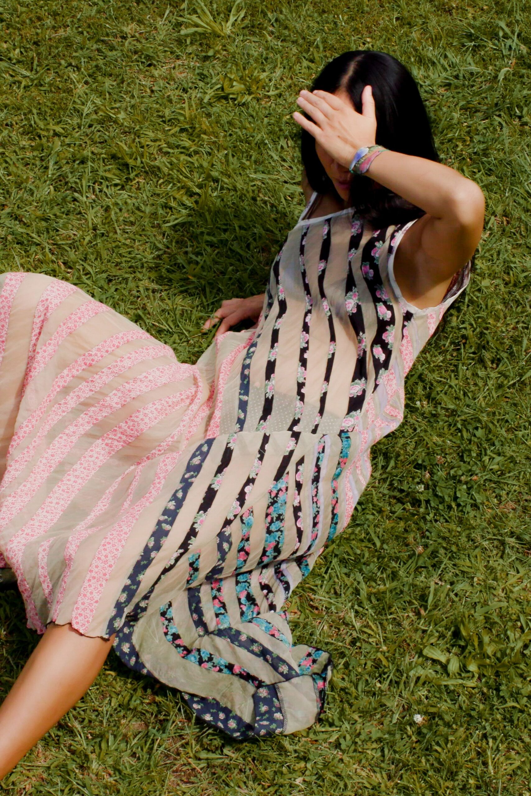 Sheer sleeveless patchwork dress, alternating gauze bands and tiny floral stripes. Pastel braided thread bracelet at the wrist. Afternoon picnic grass, squinting into sun that is stronger than expected.