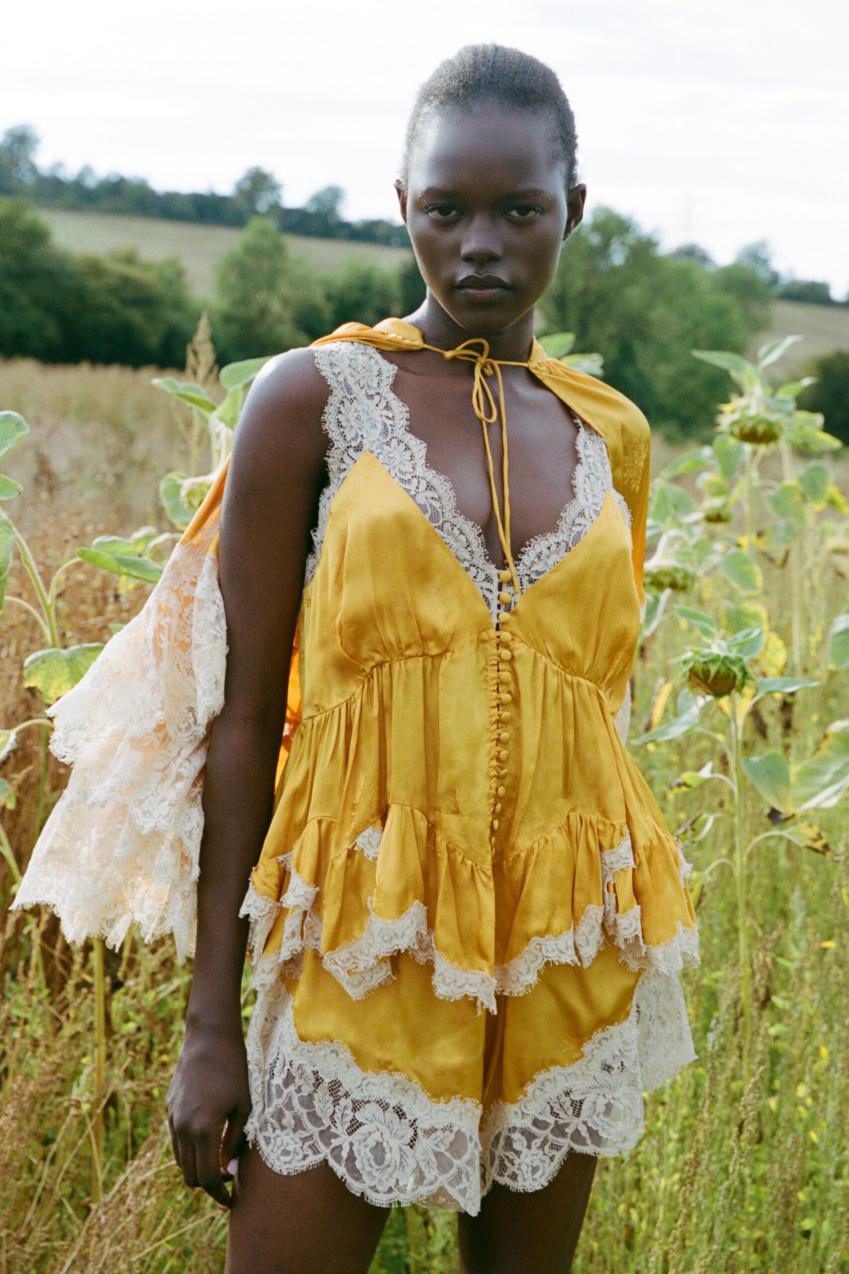 Mustard satin capelet tied at the throat with a long cord bow. Liquid satin camisole, scalloped cream lace climbing deep beside the buttons. Matching satin shorts with tiered ruffles and wide eyelash-lace hems. Sunflower field at dusk, just after taking off the wedding shoes.