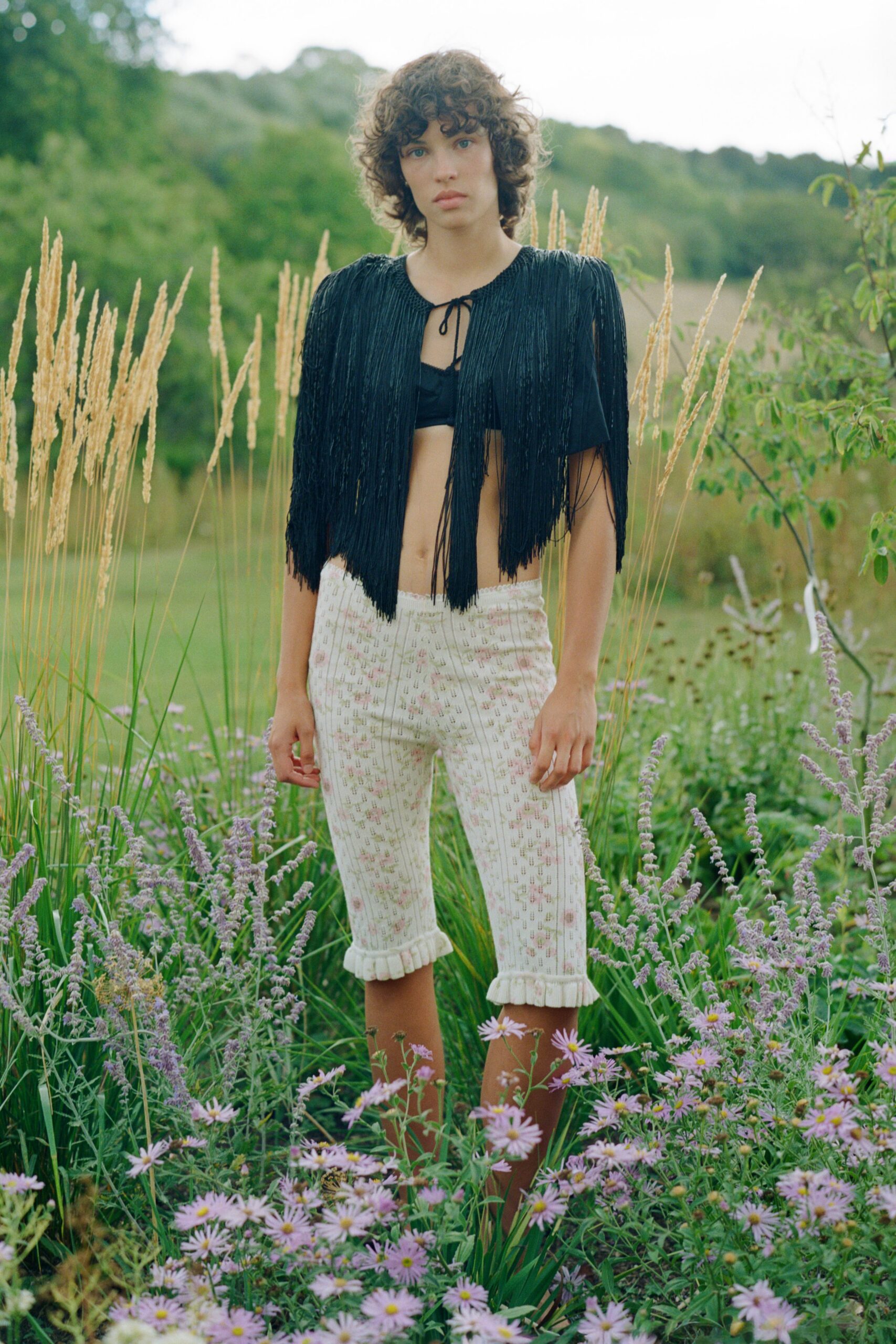 Black beaded fringe capelet, tied at the neck with thin cord. Black bandeau bralette peeking through the open front. Cream pointelle knit knickerbockers with faded pink florals and ruffled hems. Walking through waist-high asters after the field has gone quiet.
