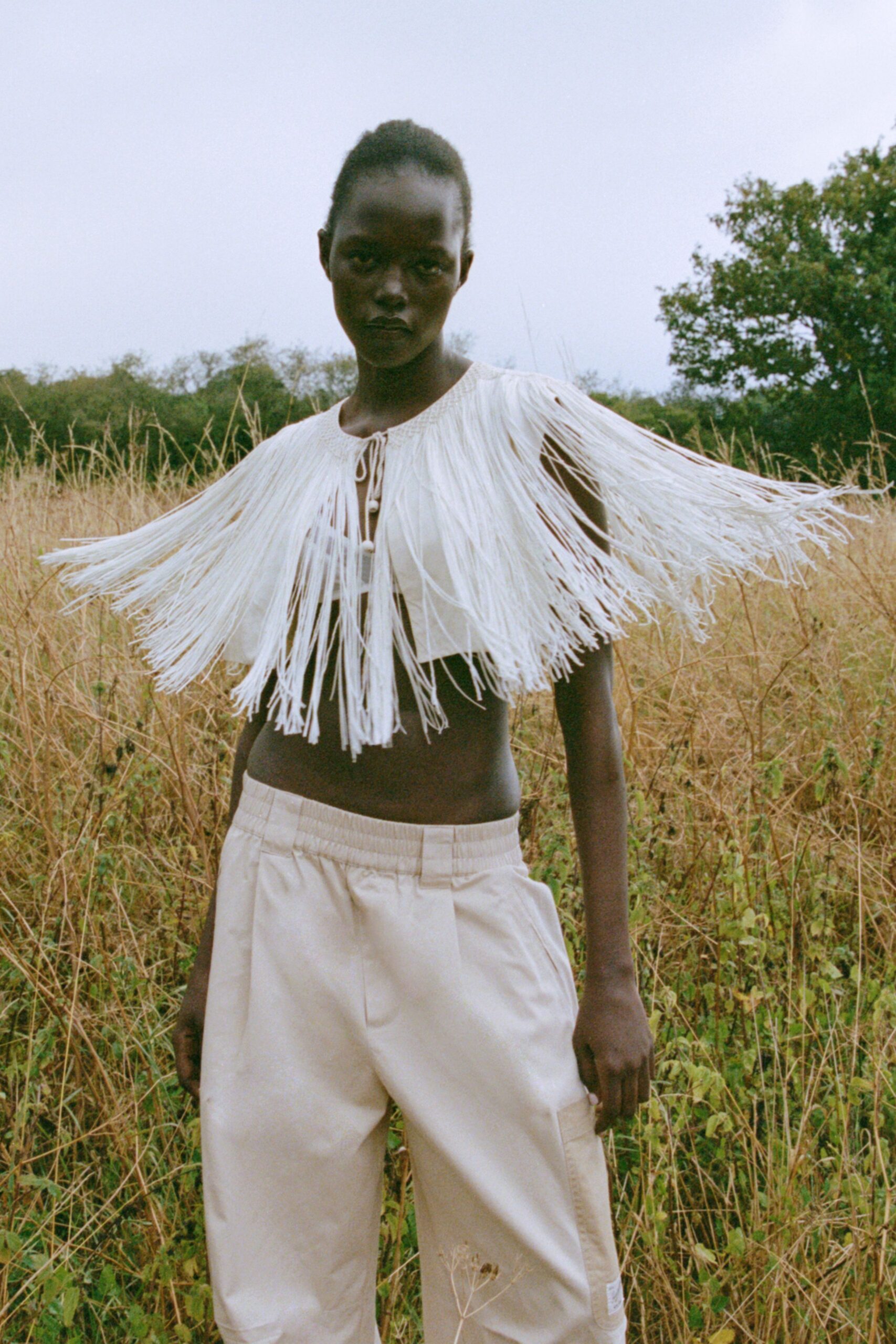 Ivory cord fringe cape, shoulder-spanning and tied with bead-ended strings. White bandeau bralette peeking beneath the swinging fringe. Cream drawstring trousers with a soft workwear rise and cargo pocket. Walking through seed-dry grass after the heat has broken.