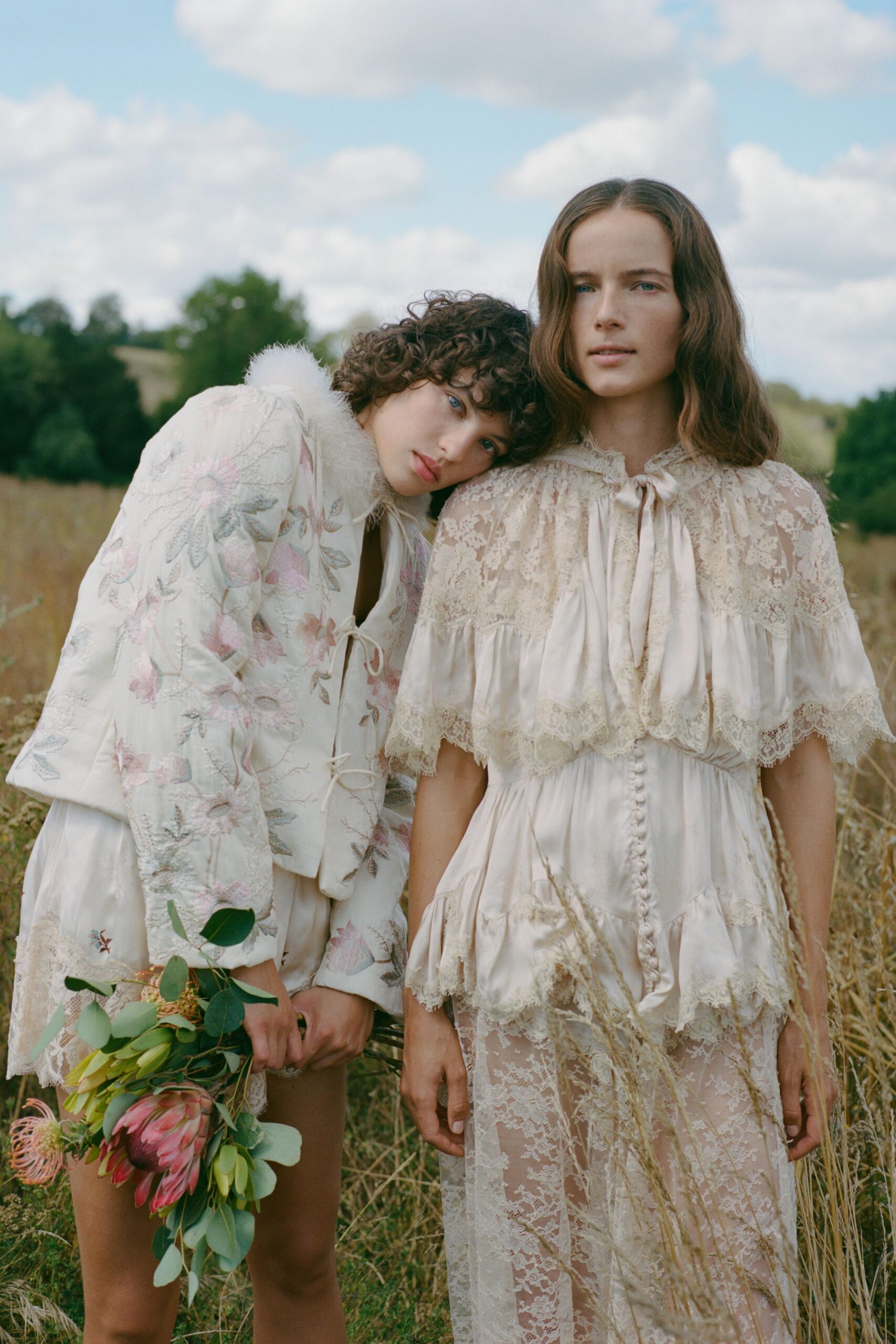 Ivory satin cape dress layered with scalloped floral lace and covered buttons. Sheer lace underlayer visible through the skirt, clouding the legs. Walking back from the meadow with seed heads caught in the hem.