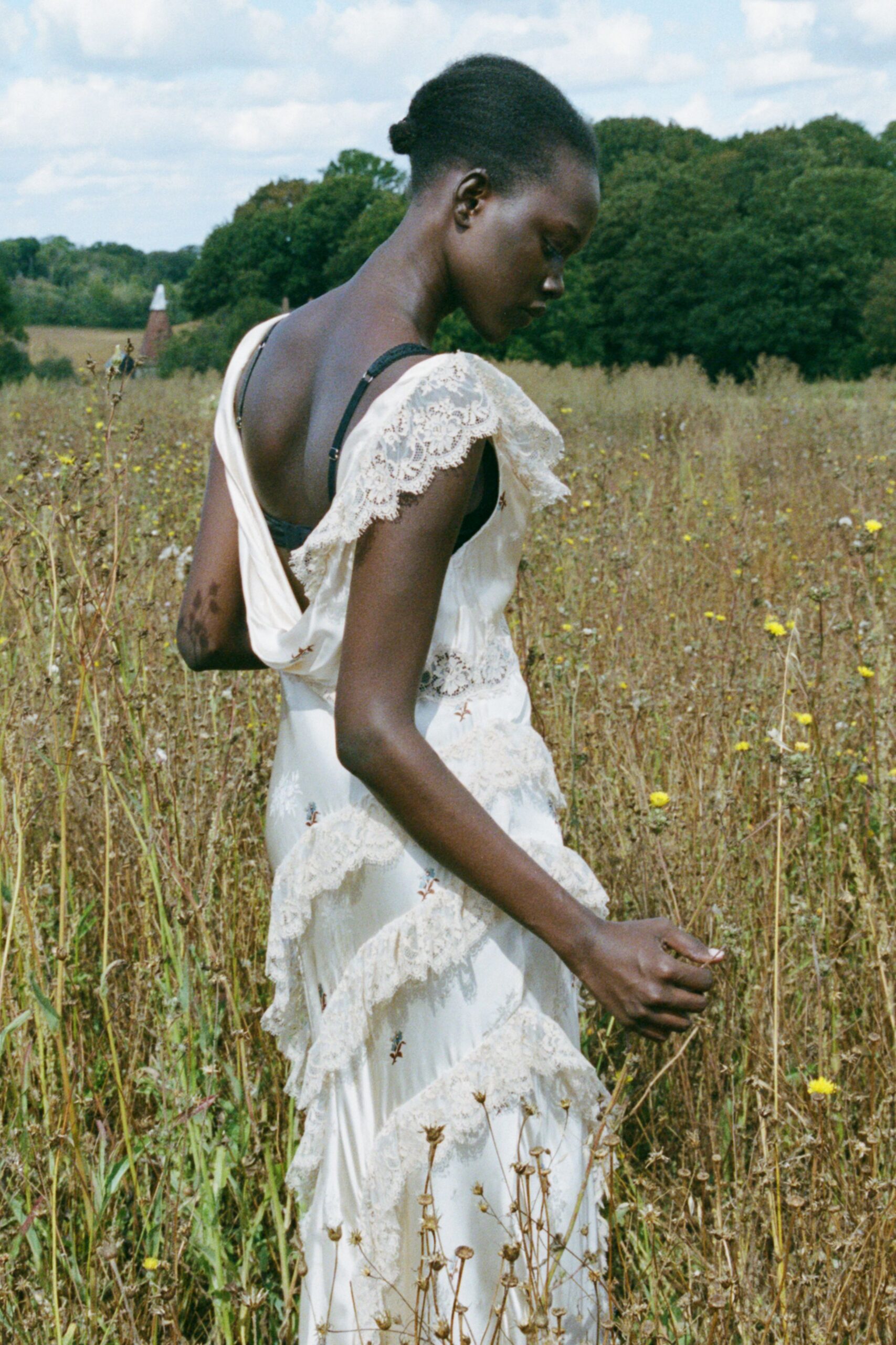 Ivory slip dress with scalloped lace flounces and a cowl-dropped back. Black bra strap cutting across the bare shoulder blade. Crossing a hay-dry field after someone else's garden wedding.