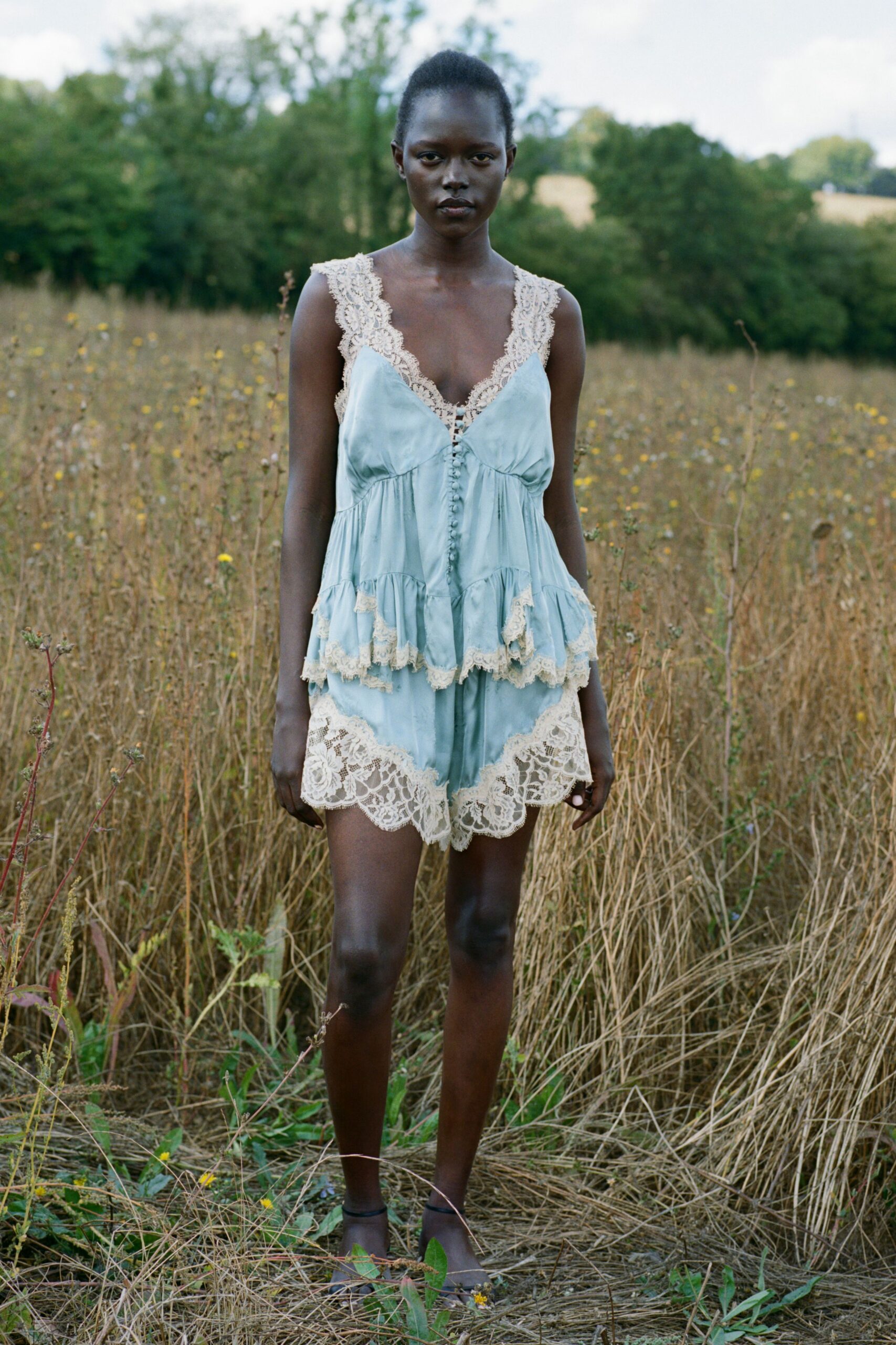 Powder-blue satin romper with scalloped cream lace straps and short hems. Tiered ruffle peplum, tiny covered buttons climbing the deep V. Black ankle-strap flats. Crossing a hayfield after rain, trying not to soak the lace.