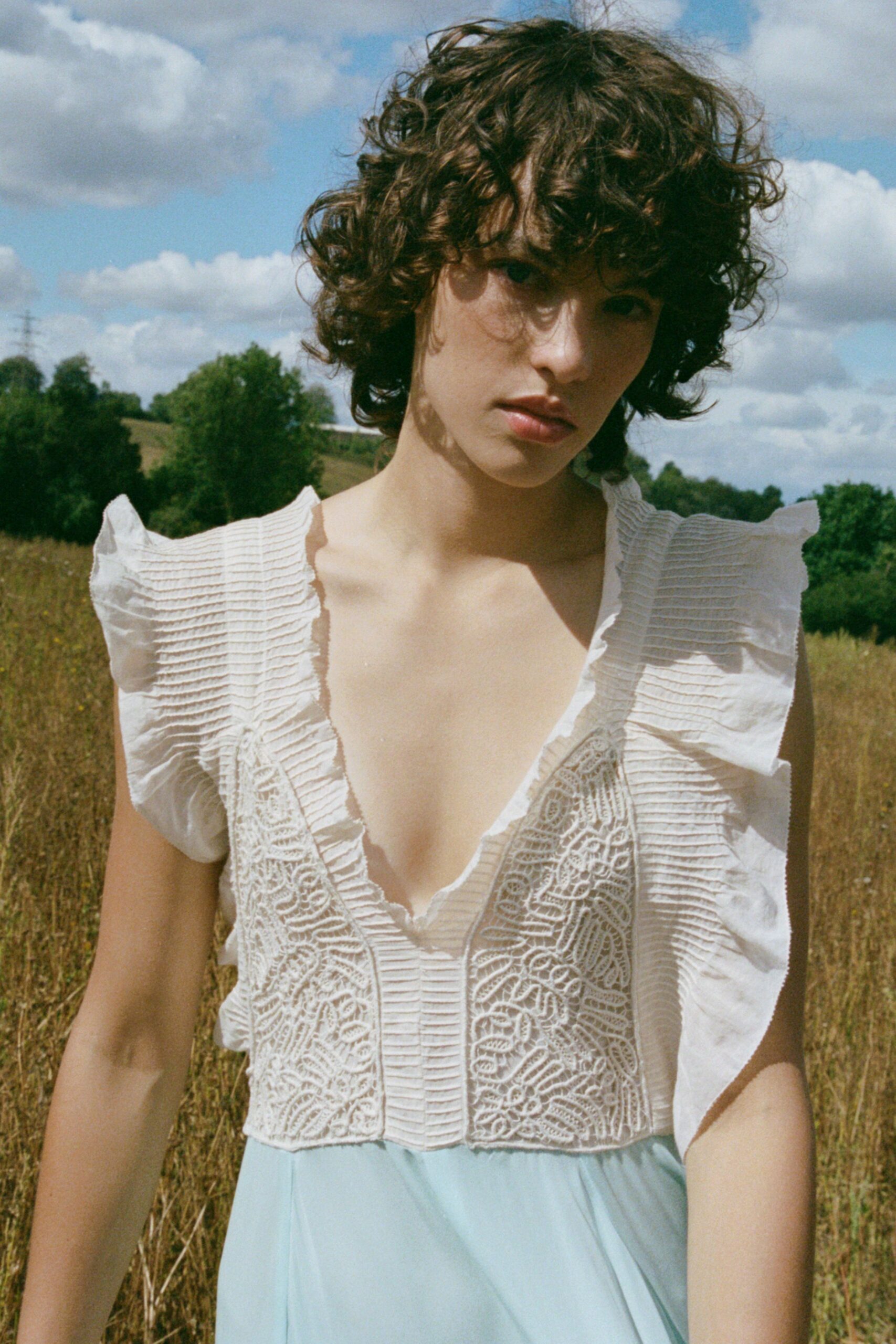 White blouse with knife-pleated ruffles and corded lace panels at the bodice. Pale blue chiffon skirt falling softly from the waist seam. Walking through waist-high grass before the storm breaks.