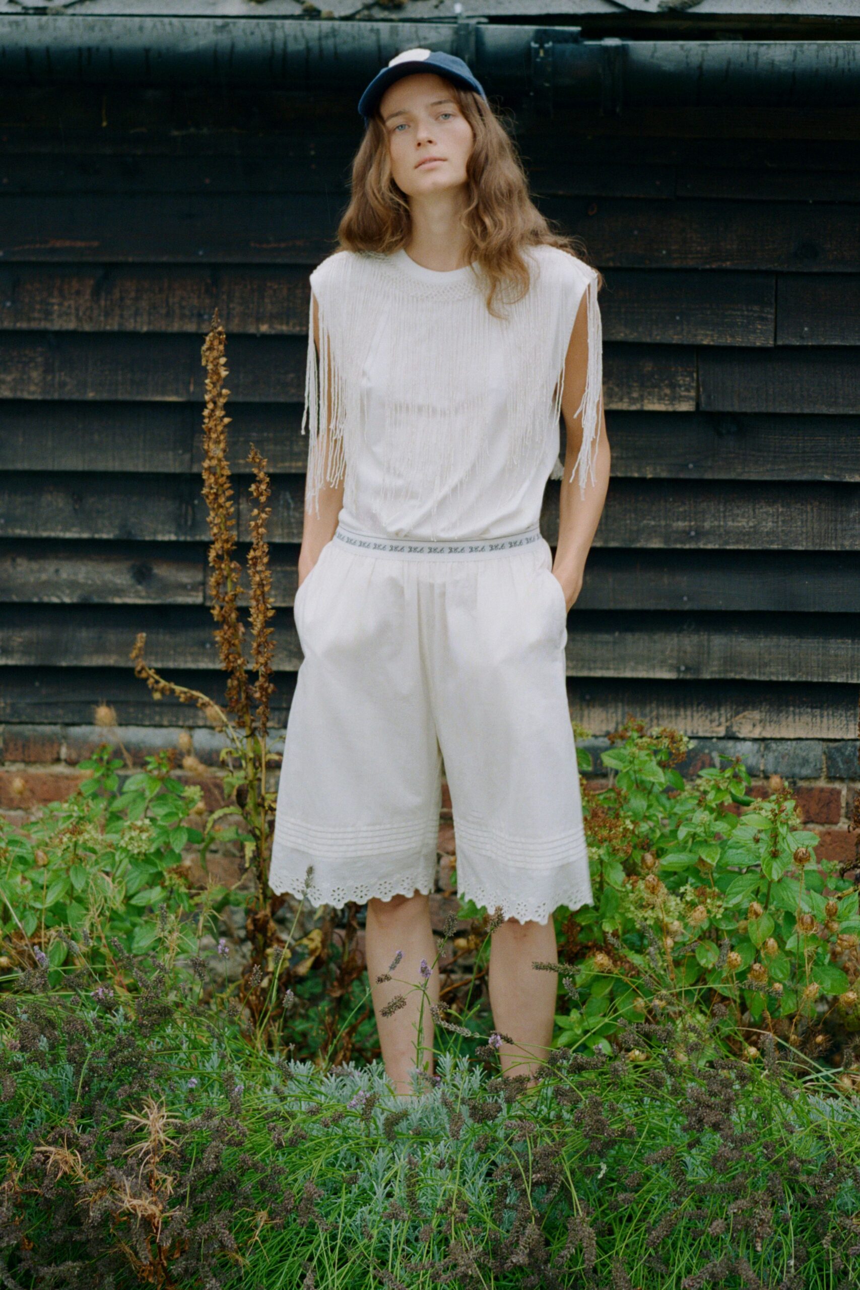 Navy baseball cap with a white front patch. White ribbed sleeveless knit, long fringe spilling from the armholes. White knee-length culottes with pintucked bands and broderie anglaise hems. Walking through the herb garden before the house wakes up.