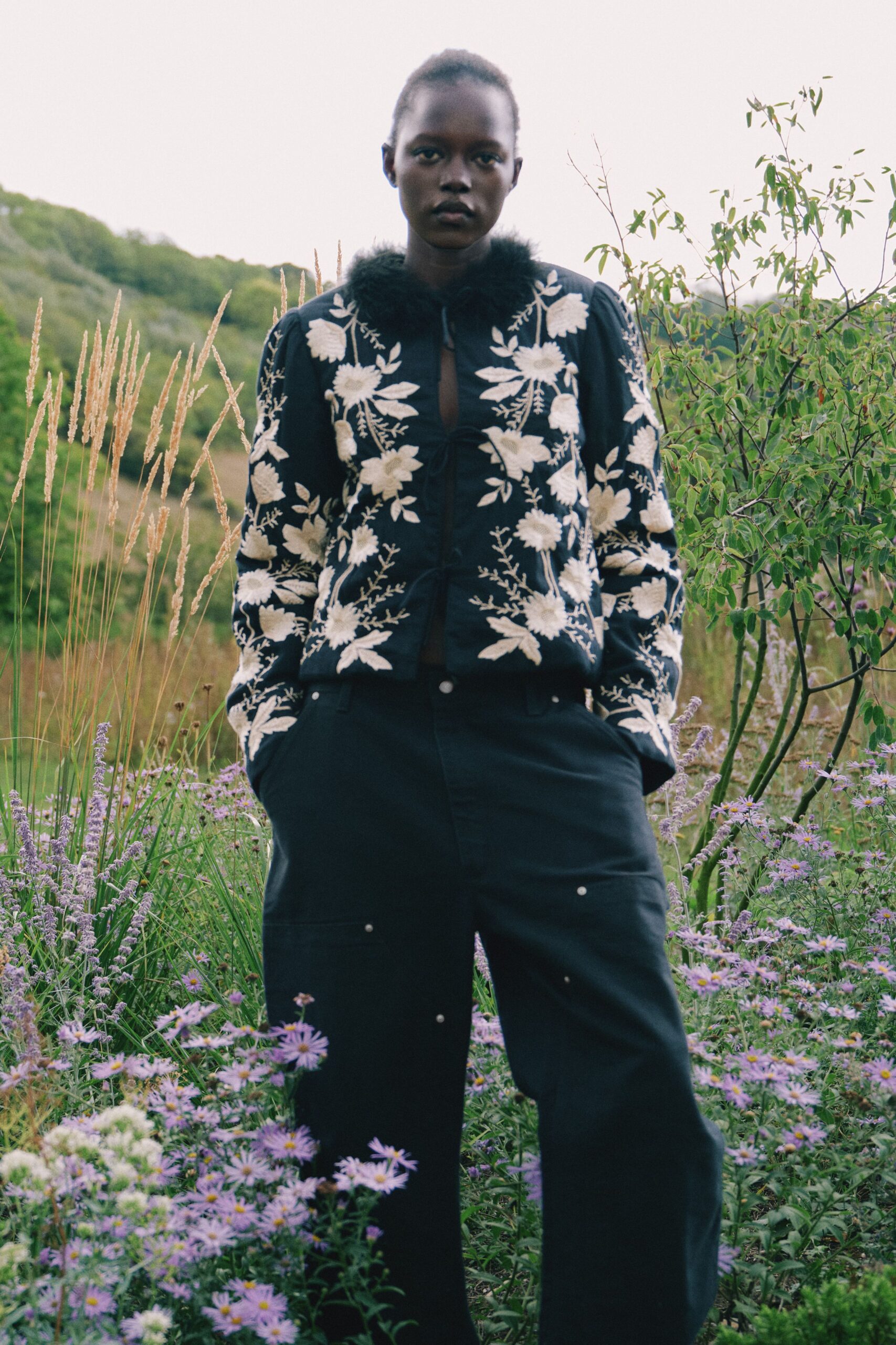 Black jacket densely embroidered with cream flowers, ringed by a fluffy fur collar. Black wide-leg trousers with silver stud dots and deep patch pockets. Walking through an overgrown garden after rain, asters brushing both shins.