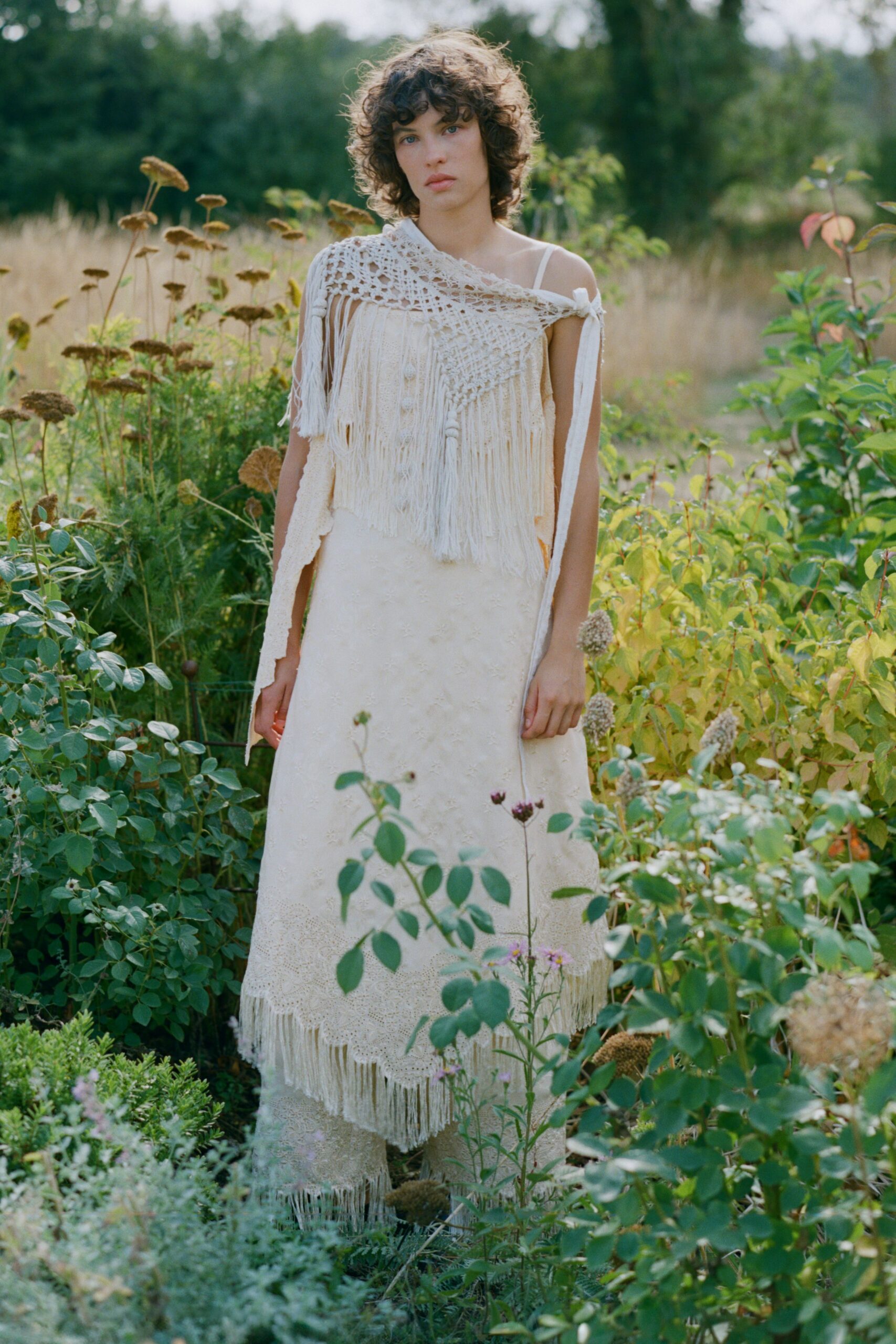 Openwork cream crochet draped off one shoulder, knotted with dense fringe. Ivory slip dress with tiny tonal floral embroidery and a lace-fringed hem. Cream lace trousers ending in another swaying fringe band. In the herb garden after rain, cutting stems with nowhere urgent to be.