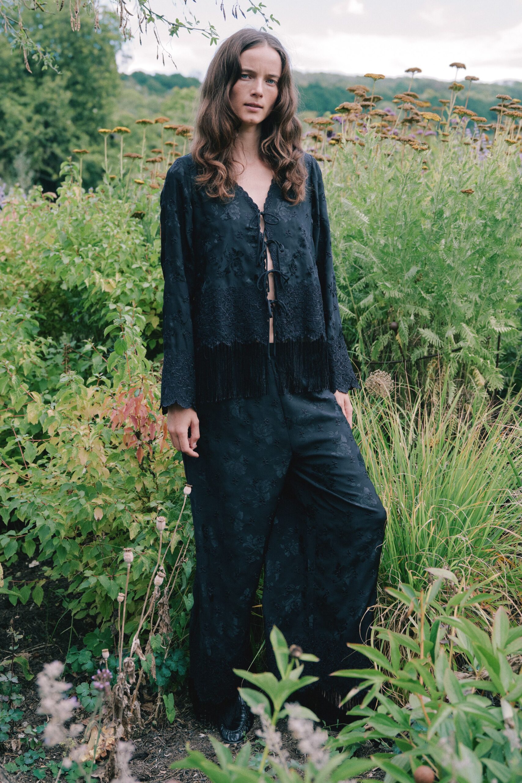 Black floral jacquard blouse, tied open with long fringe at the hem. Matching wide-leg satin trousers, scattered with tonal flower motifs. Black leather toes barely peeking beneath the break. Walking the herb garden after rain, stems brushing both ankles.