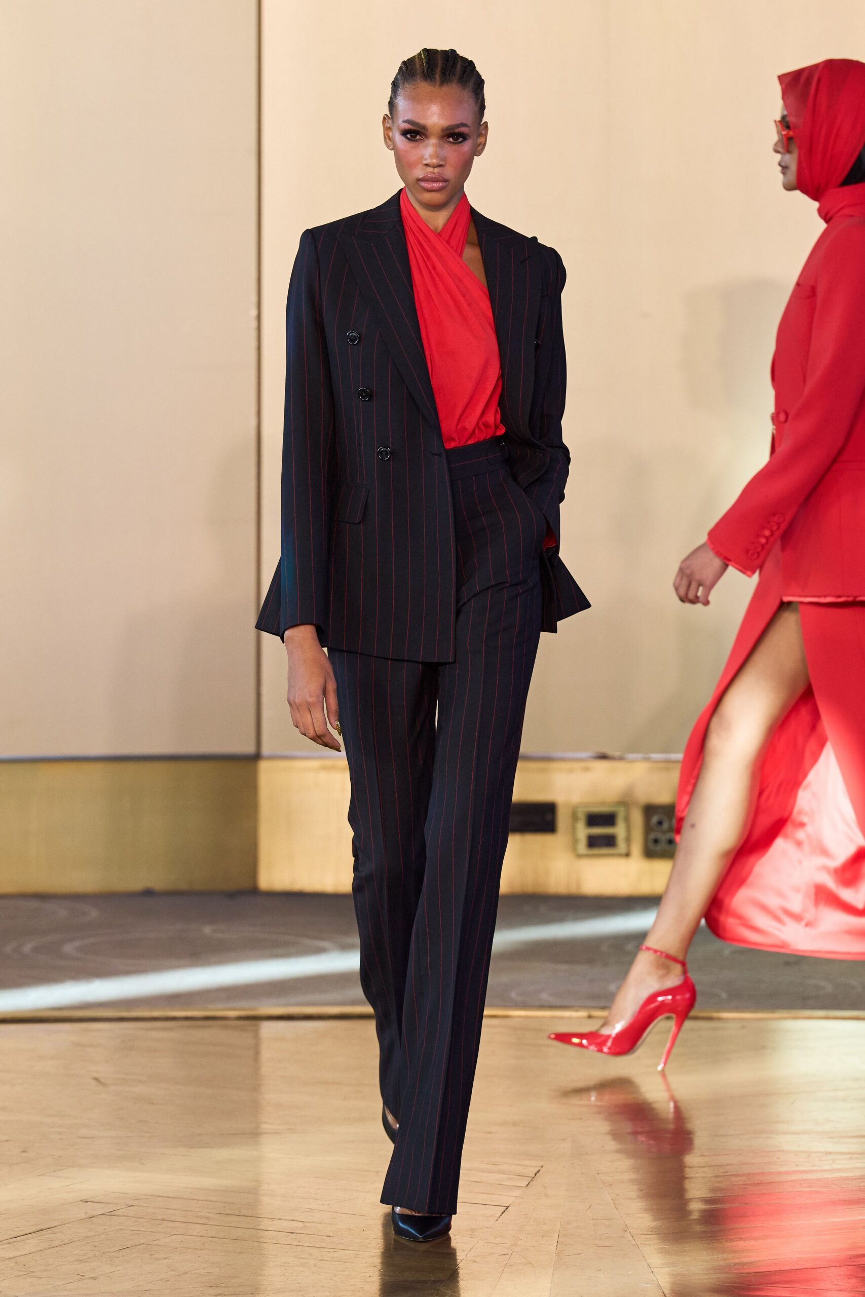 Black double-breasted blazer traced with narrow oxblood pinstripes. Scarlet jersey top crossing high at the neck in soft folds. Matching pinstripe trousers cut straight and long over the shoe. Black satin pointed heels. For taking the corner office key after everyone else has gone home.