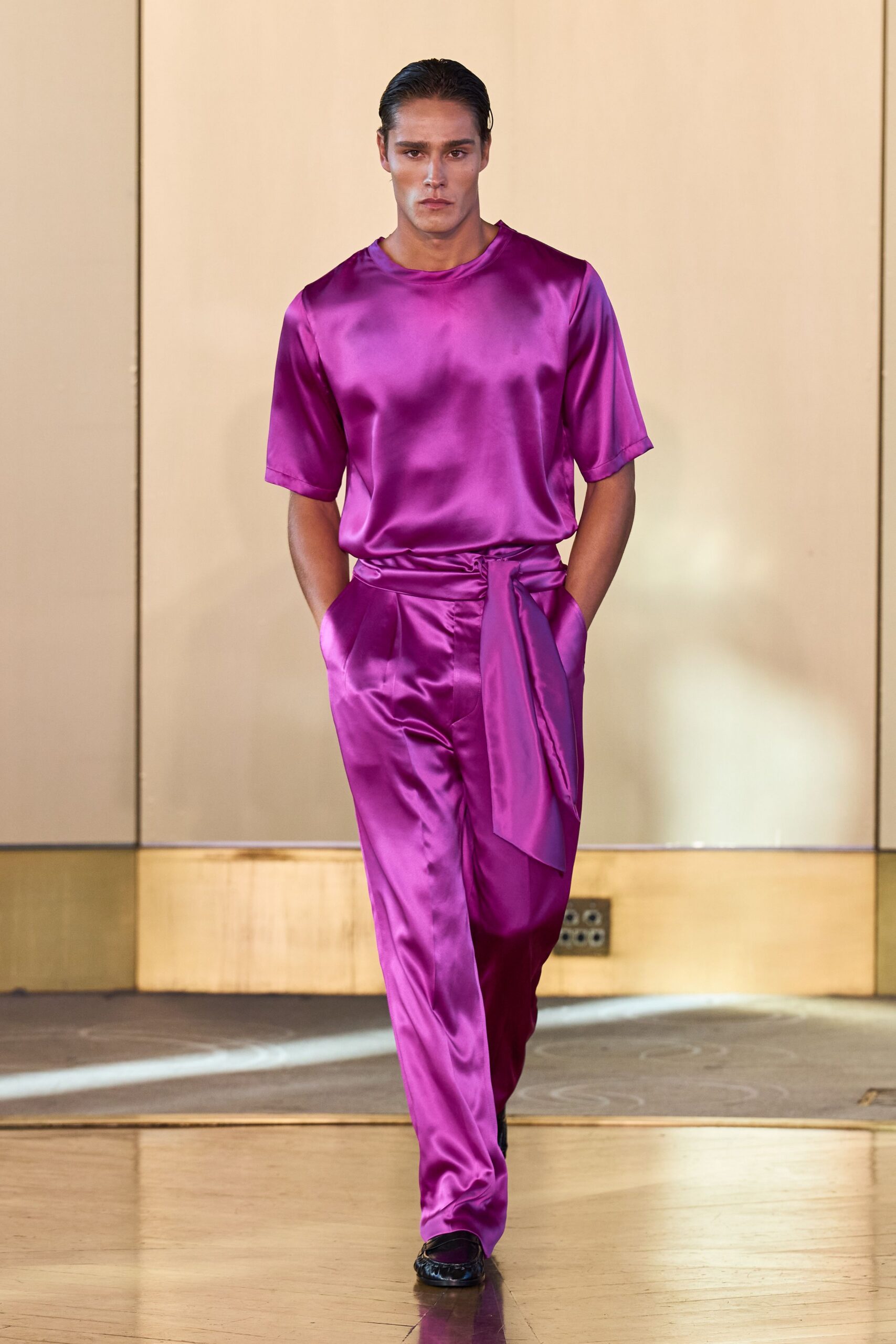 Fuchsia satin top with a clean round neck and elbow sleeves. Matching satin trousers, high-waisted with a wide sash knotted off-center. Black loafers peeking from the pooling hem. Hotel corridor at noon, carrying flowers back from someone else's wedding.