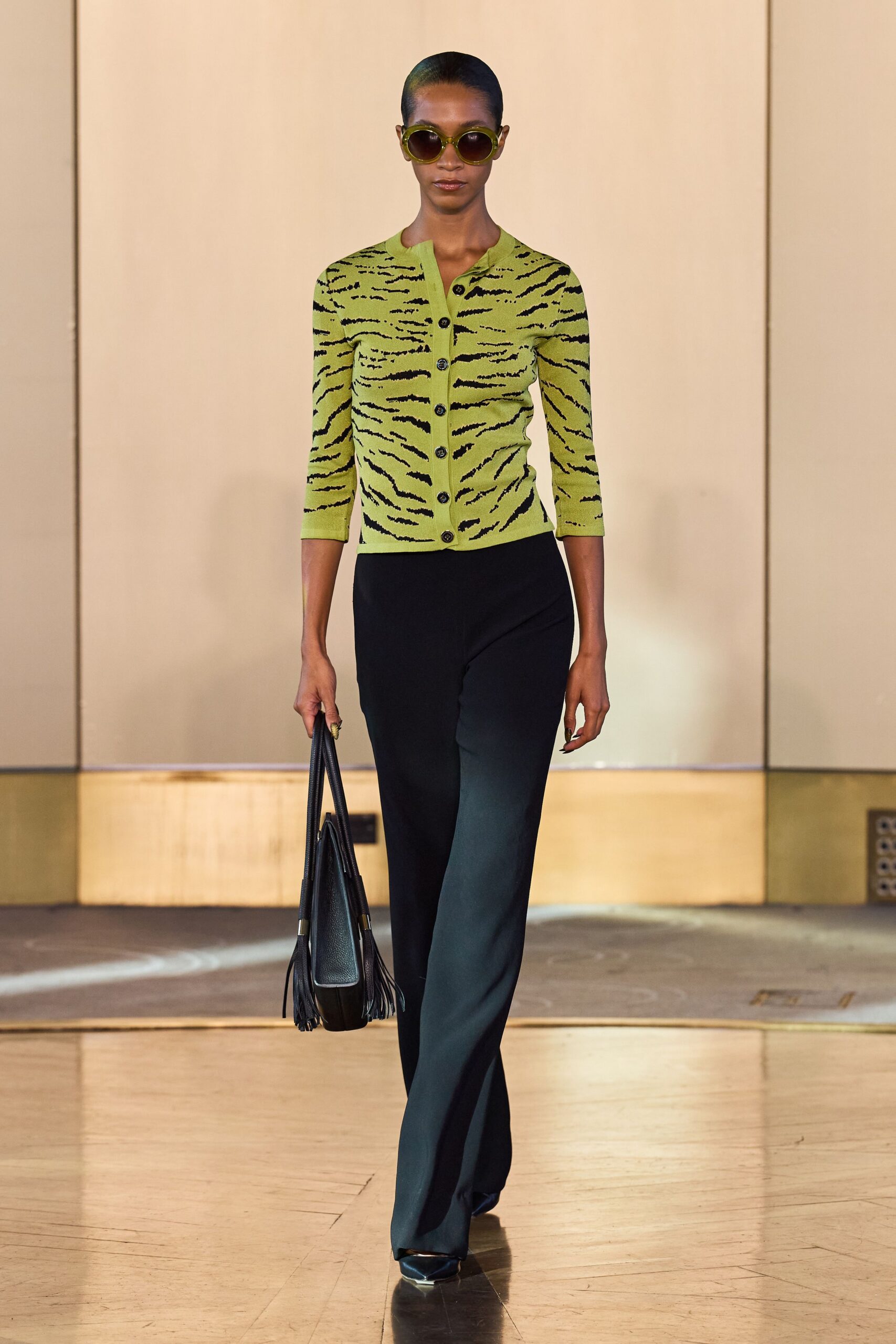 Round olive frames with dark lenses. Chartreuse knit cardigan striped like tiger fur, third button catching light. Black floor-grazing trousers with a pressed front crease. Black pebbled leather tote with long fringe at the sides. Pointed black leather pumps. Walking into a hotel lobby before noon with your sunglasses still on.