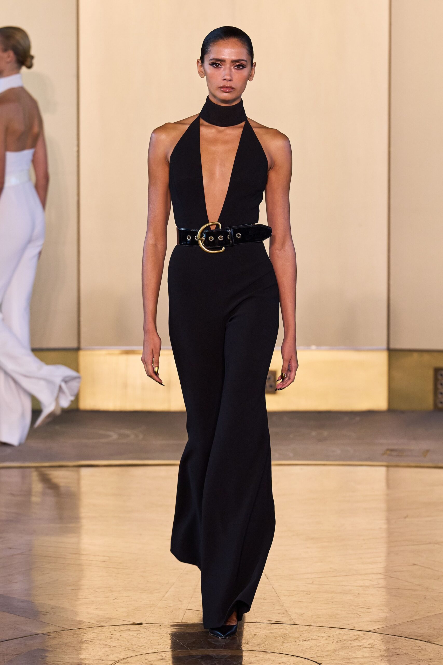 Black halter jumpsuit with a plunging V and built-in choker collar. Wide patent belt with a brushed gold buckle and eyelets. Black pointed pumps, mostly swallowed by the flared hem. Hotel ballroom after midnight, carrying yourself like the room already cleared.