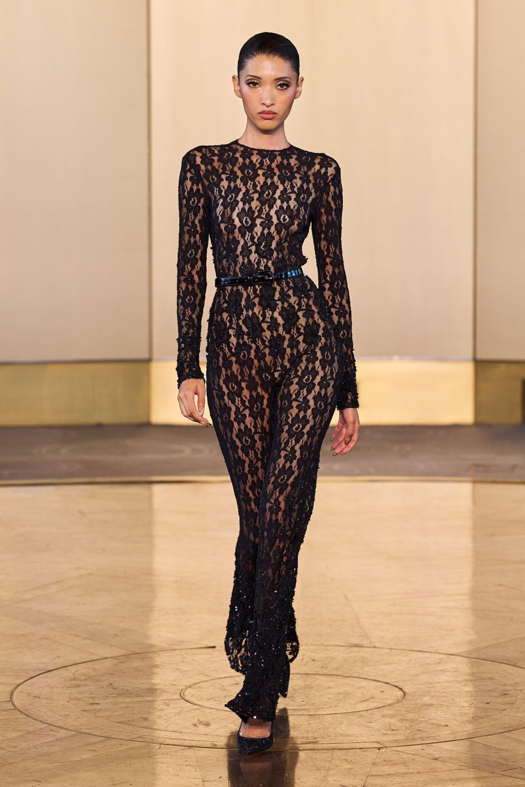 Sheer black floral lace catsuit over nude lining, crew neck and long sleeves. Narrow patent belt, square buckle catching the light at center waist. Black glitter pumps with a rounded vamp. For crossing a hotel ballroom after midnight when the carpet still hums.