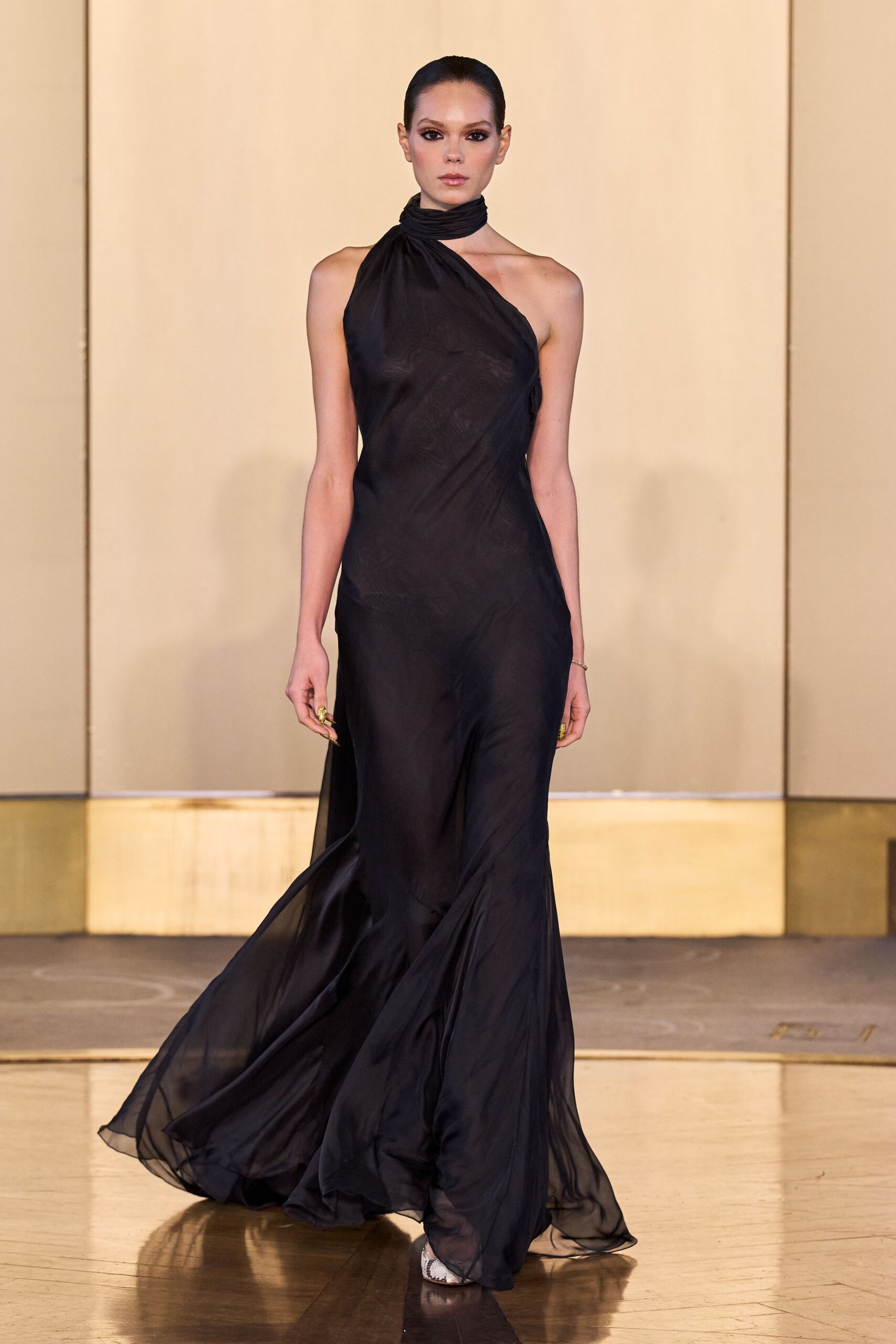 Black chiffon gown cut on the bias, one-shoulder drape twisting into a neck wrap. Chunky gold rings catching at both hands. Python pointed heels flashing under the pooling hem. Whatever you wear to cross a hotel lobby after midnight without breaking stride.